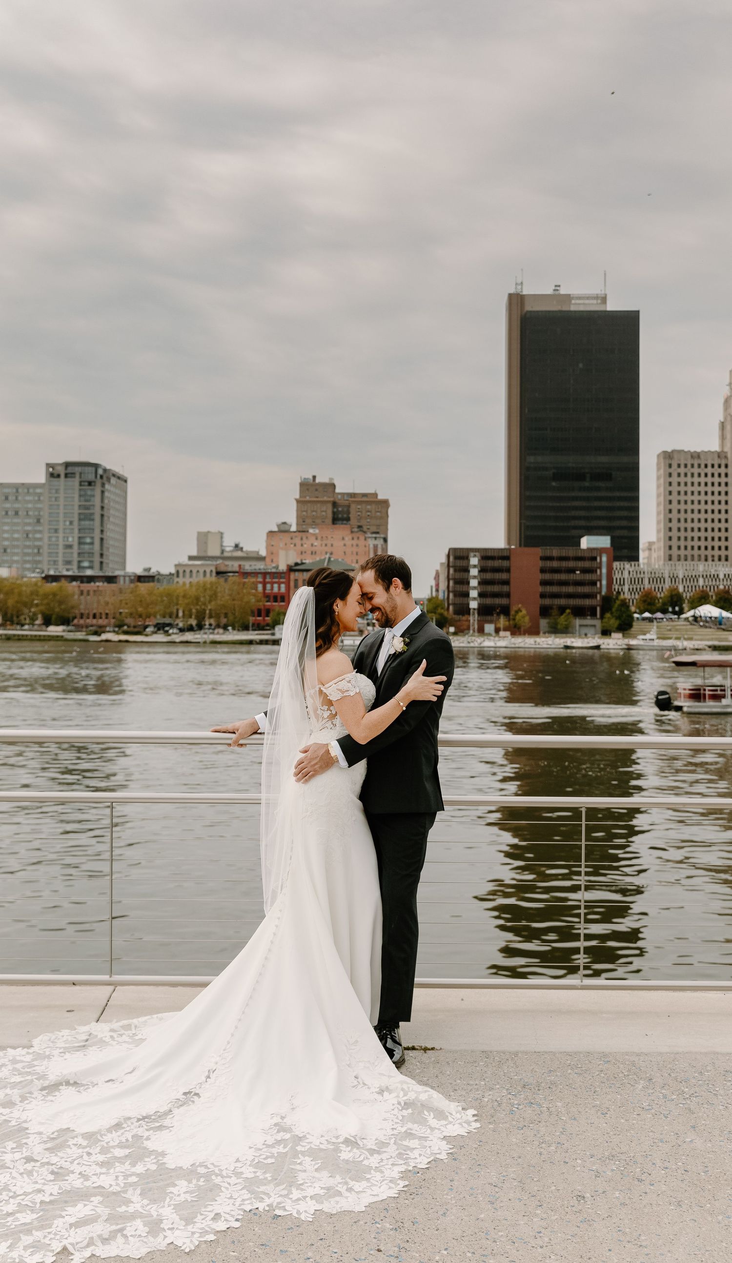 Wedding couple shares romantic dance on waterfront terrace with city skyline and river in background.
