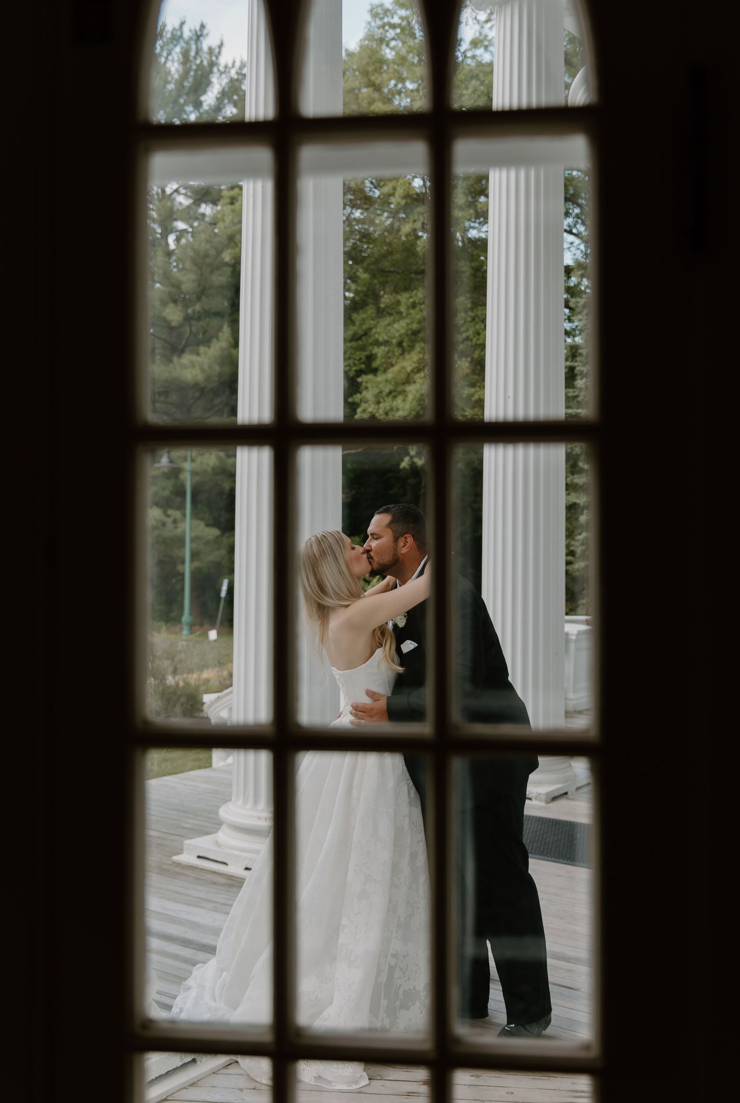 Bride and groom kiss passionately on a columned portico, captured through a classic divided-light window frame.