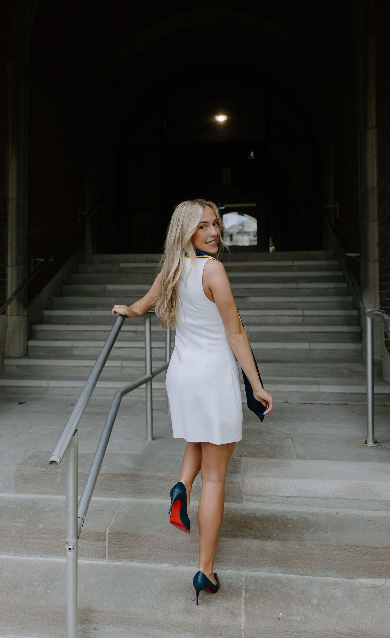 Blonde woman in white mini dress and red-soled heels ascending stone stairs, glancing back over her shoulder.