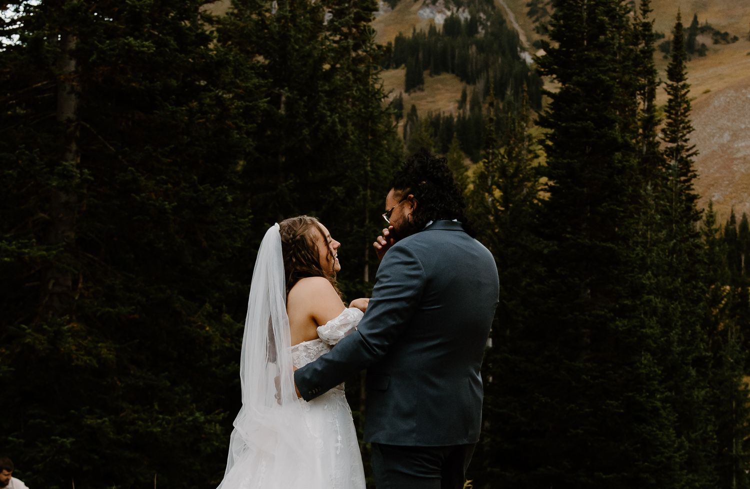 A couple in wedding attire dances together surrounded by tall evergreen trees at sunset.