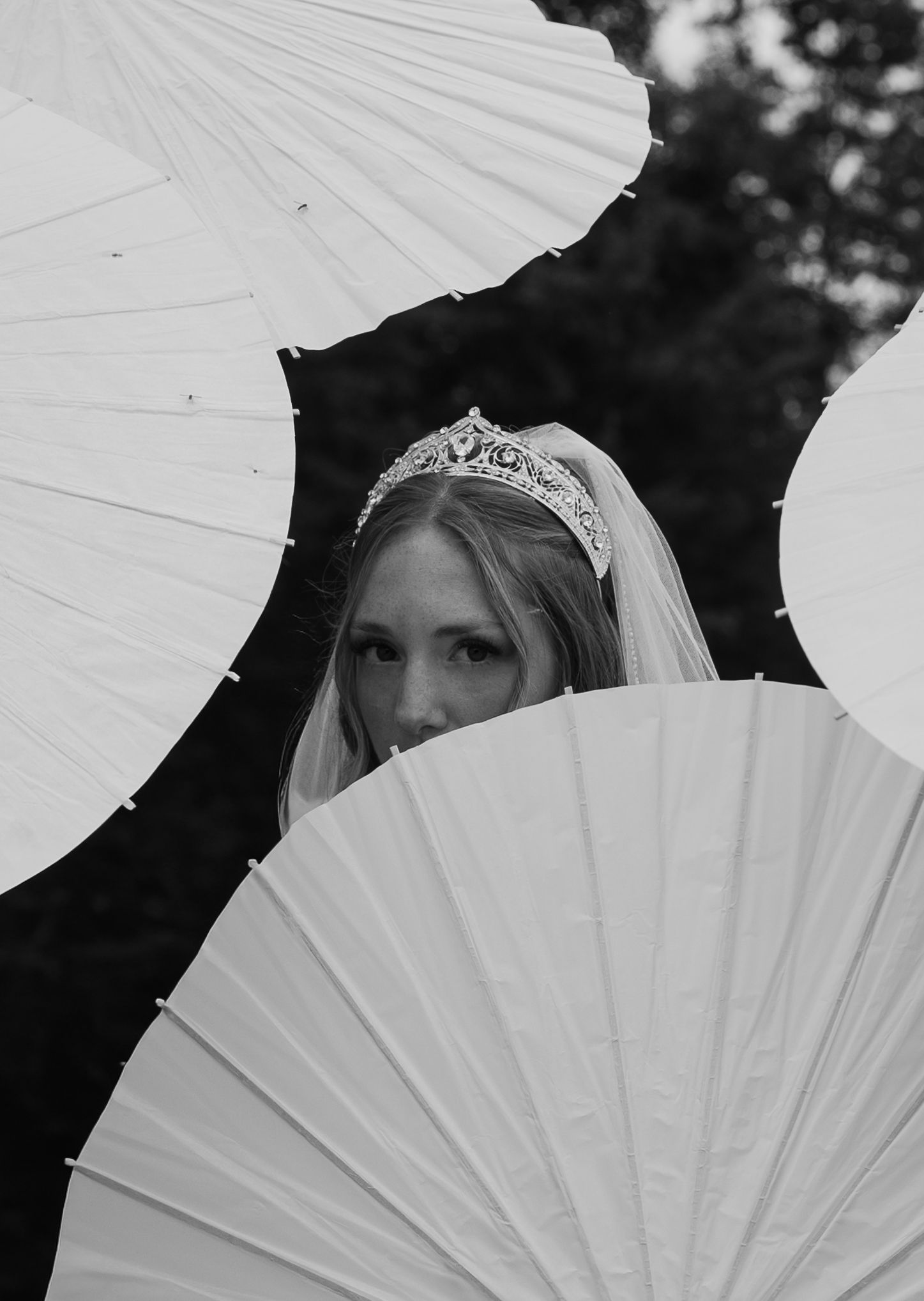 Black and white photo of a bride wearing a tiara and veil, partially hidden behind white paper parasols.