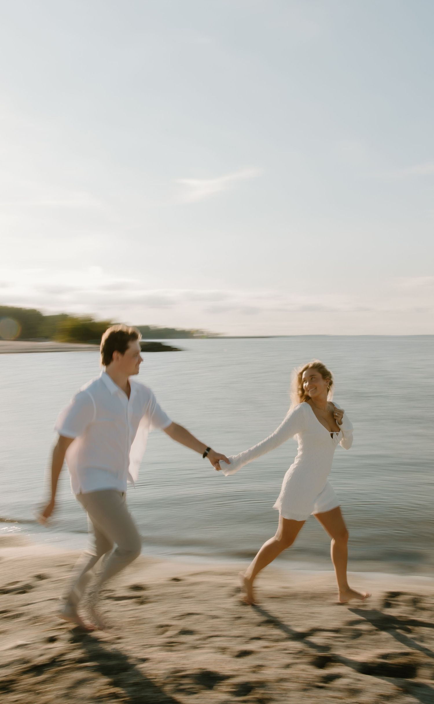 A couple in white outfits holds hands while running joyfully along a sandy beach at sunset.