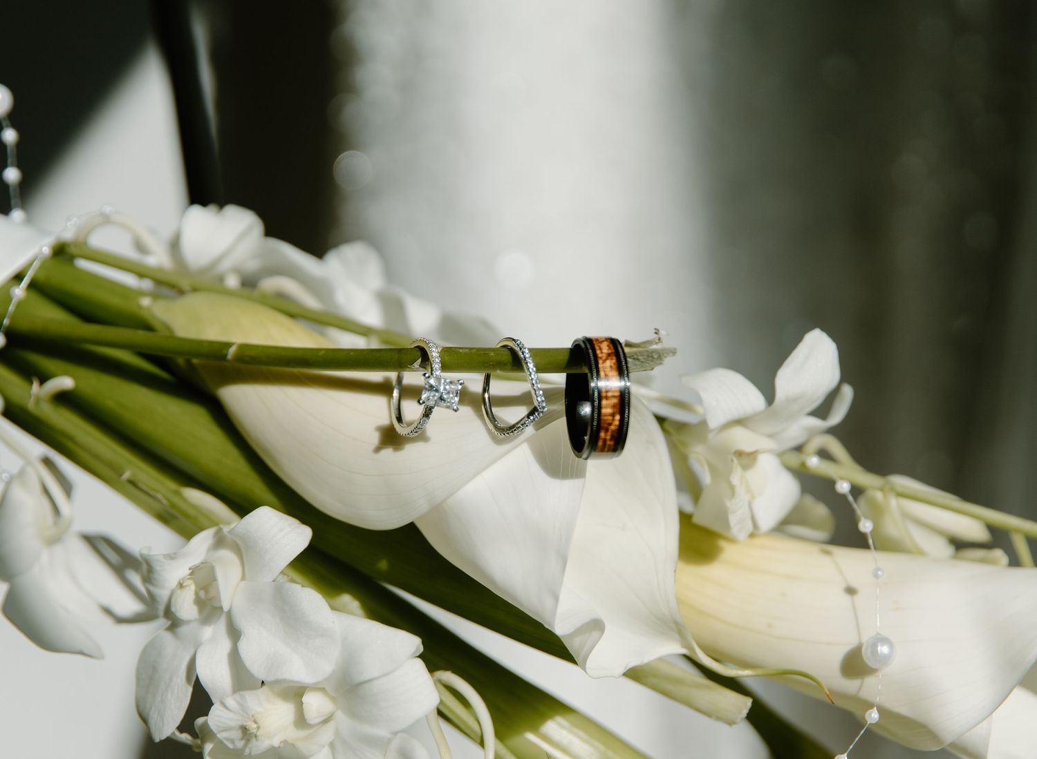 Wedding rings rest elegantly on white lily petals with green stems in a romantic macro shot.