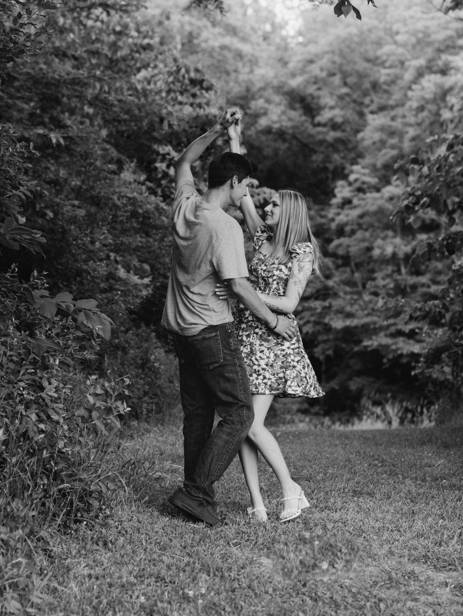 A black and white photo of a couple dancing together in a wooded outdoor setting.