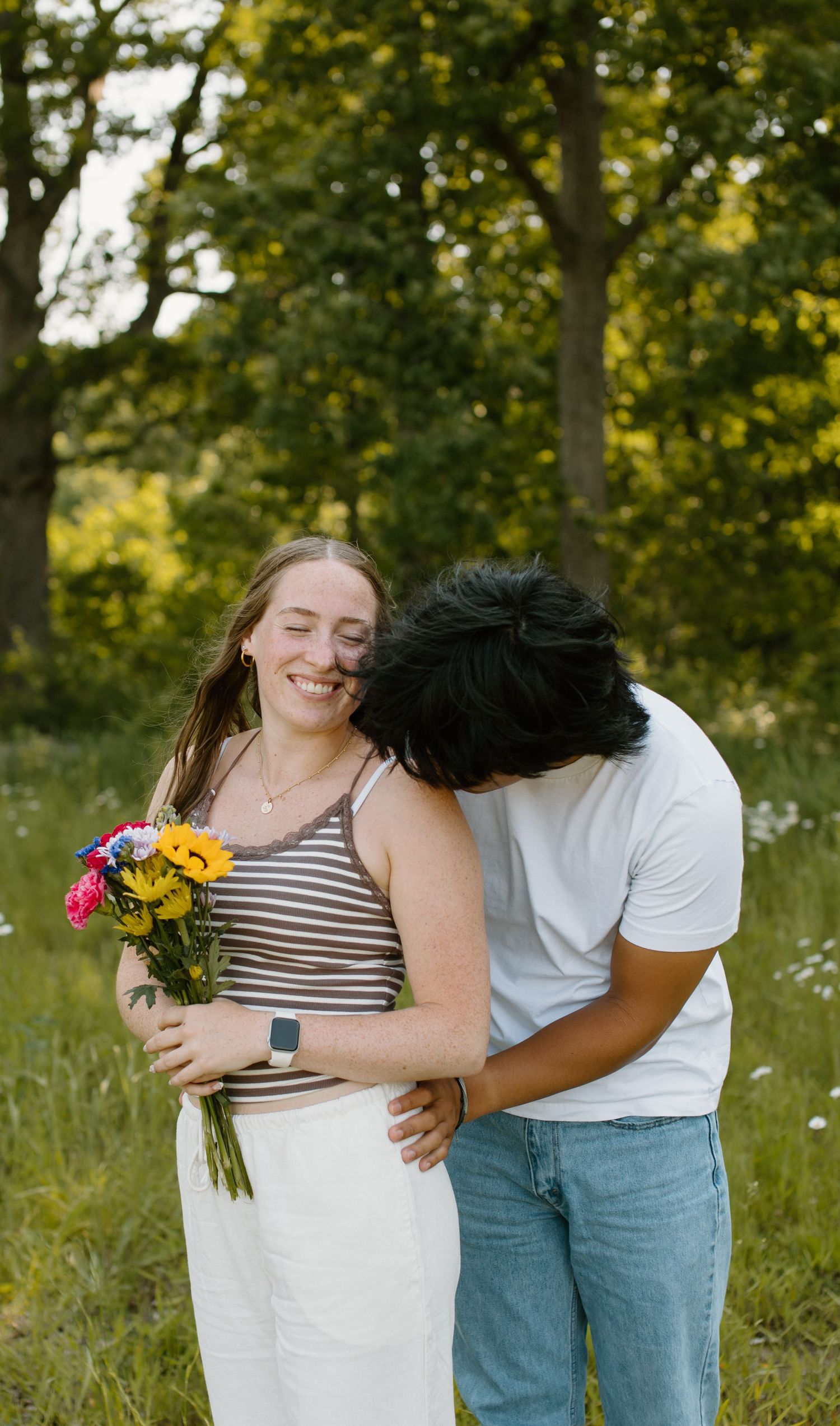 A couple shares a tender moment outdoors while holding a colorful bouquet of sunflowers and pink flowers in a natural setting.