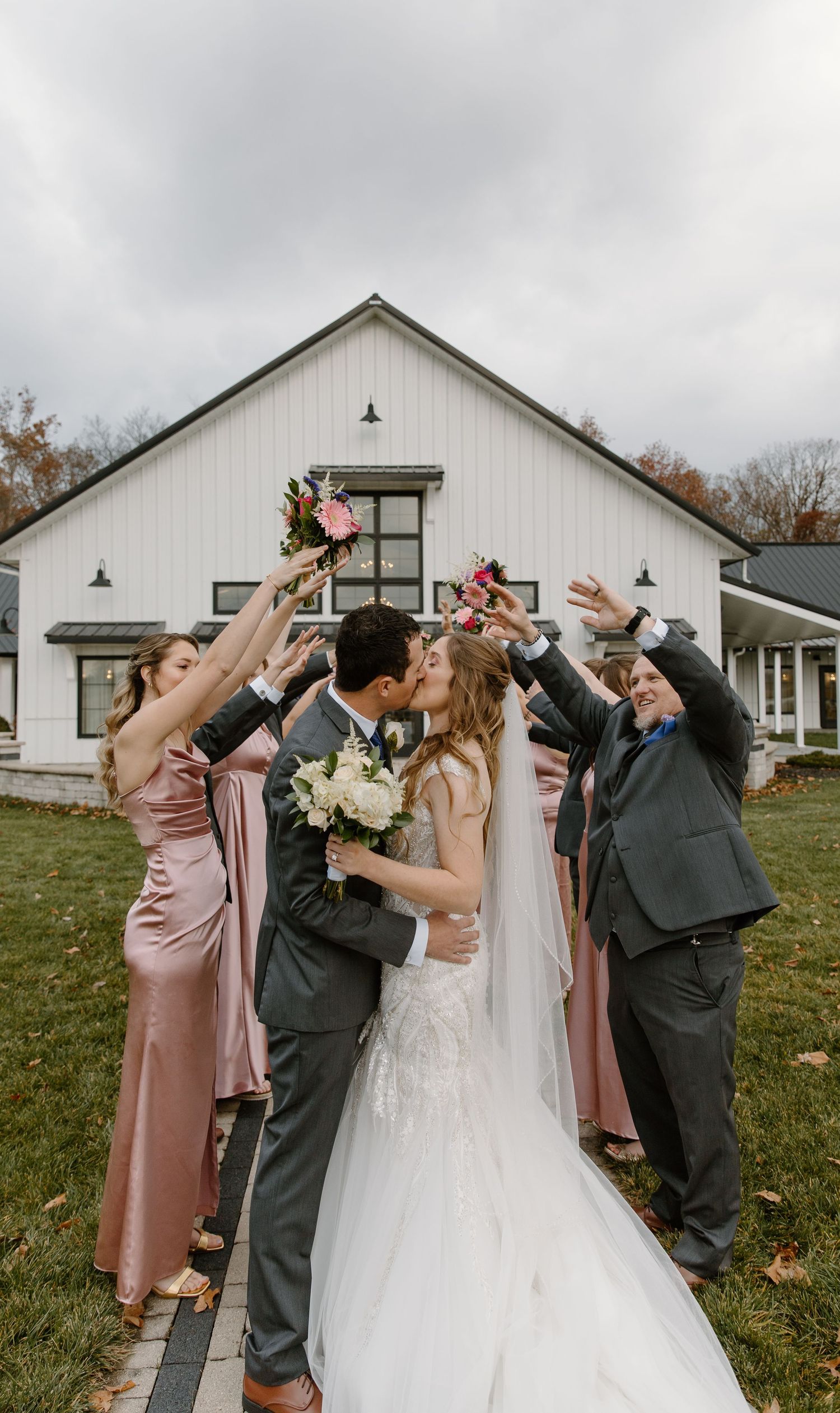 Wedding party celebrates with champagne toast in front of white barn on cloudy autumn day.
