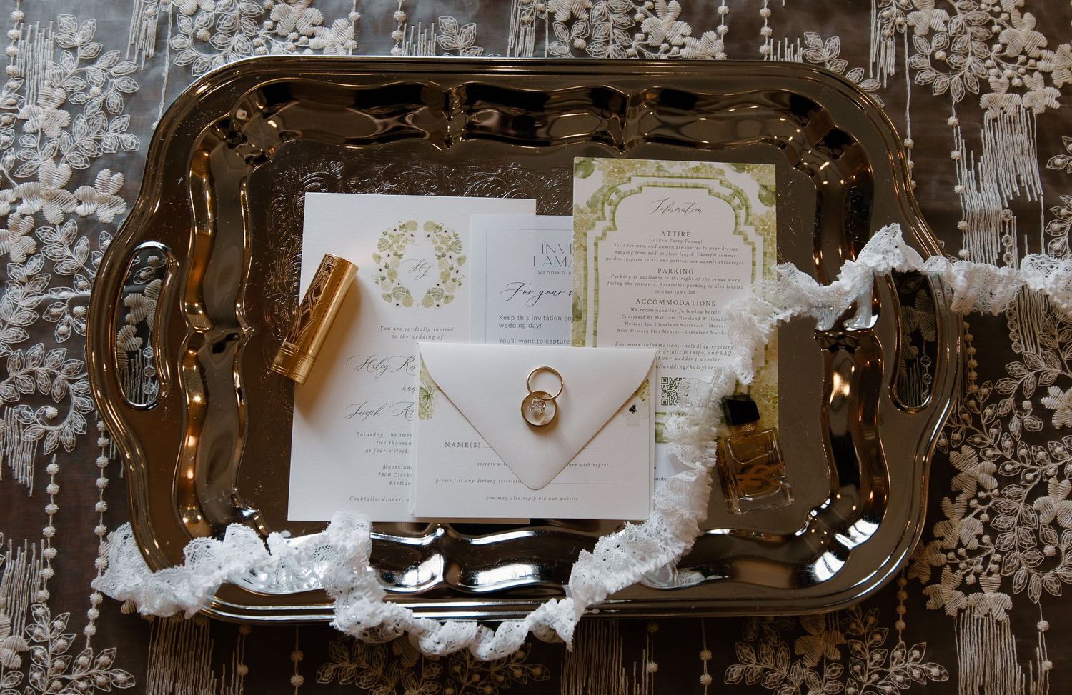 Elegant wedding invitation suite with gold wreath design displayed on ornate silver tray with rings and ribbon accents.