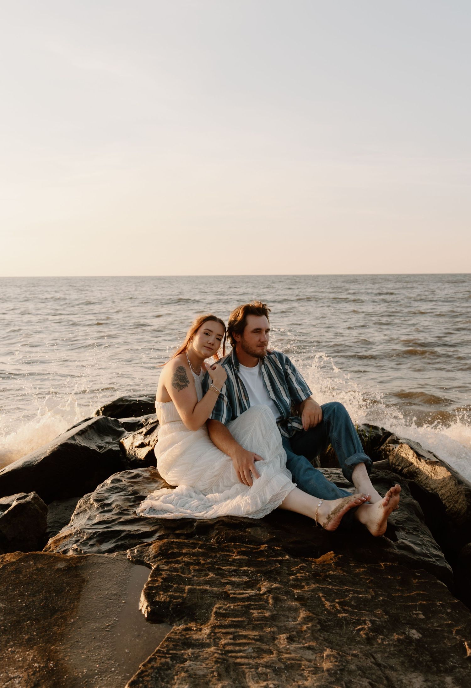 A couple sits on rocky shoreline at golden hour, waves crashing behind them as woman leans on mans shoulder.