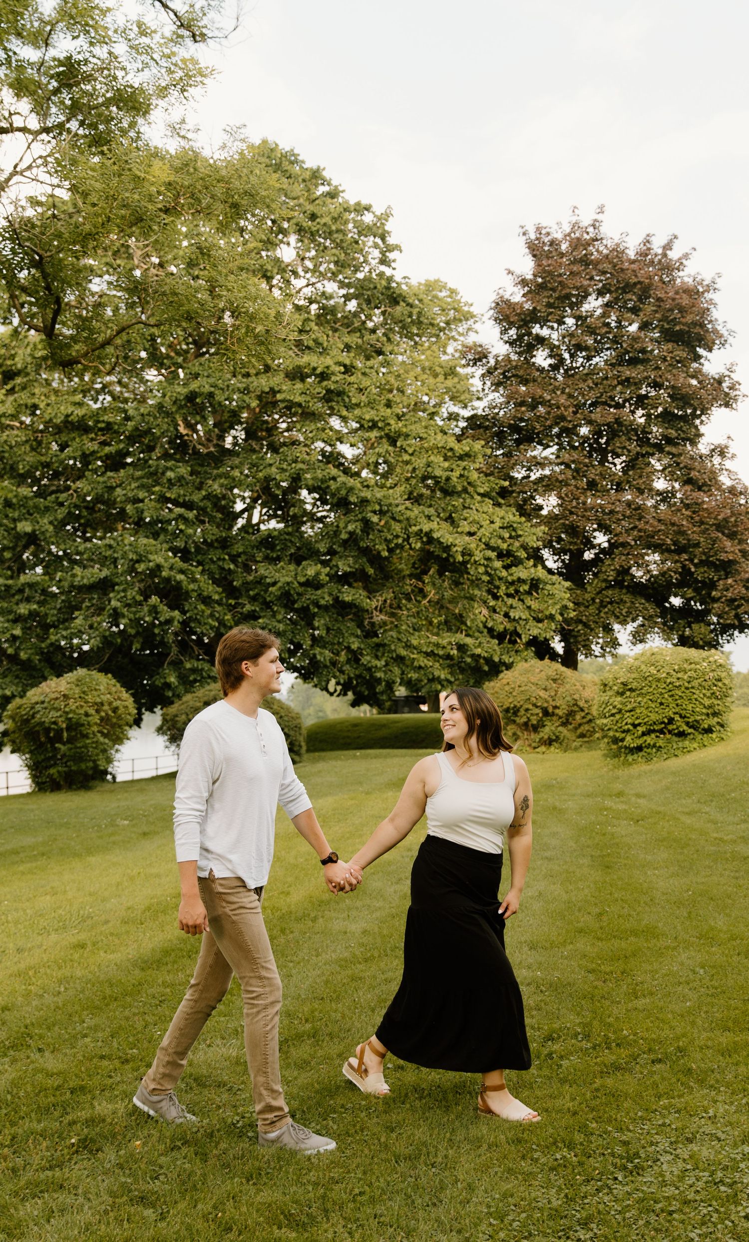Two people hold hands while walking on a lush green lawn with mature trees in the background on a summer day.