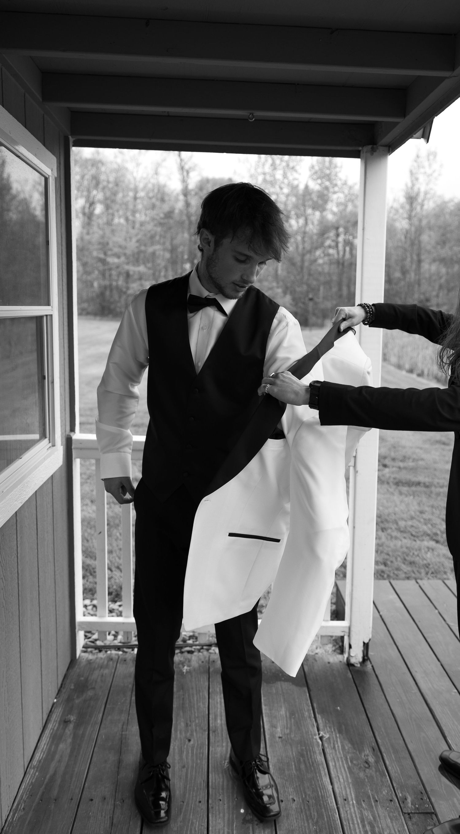 Young man in tuxedo vest and bow tie being helped into white jacket on wooden porch, black and white photo.