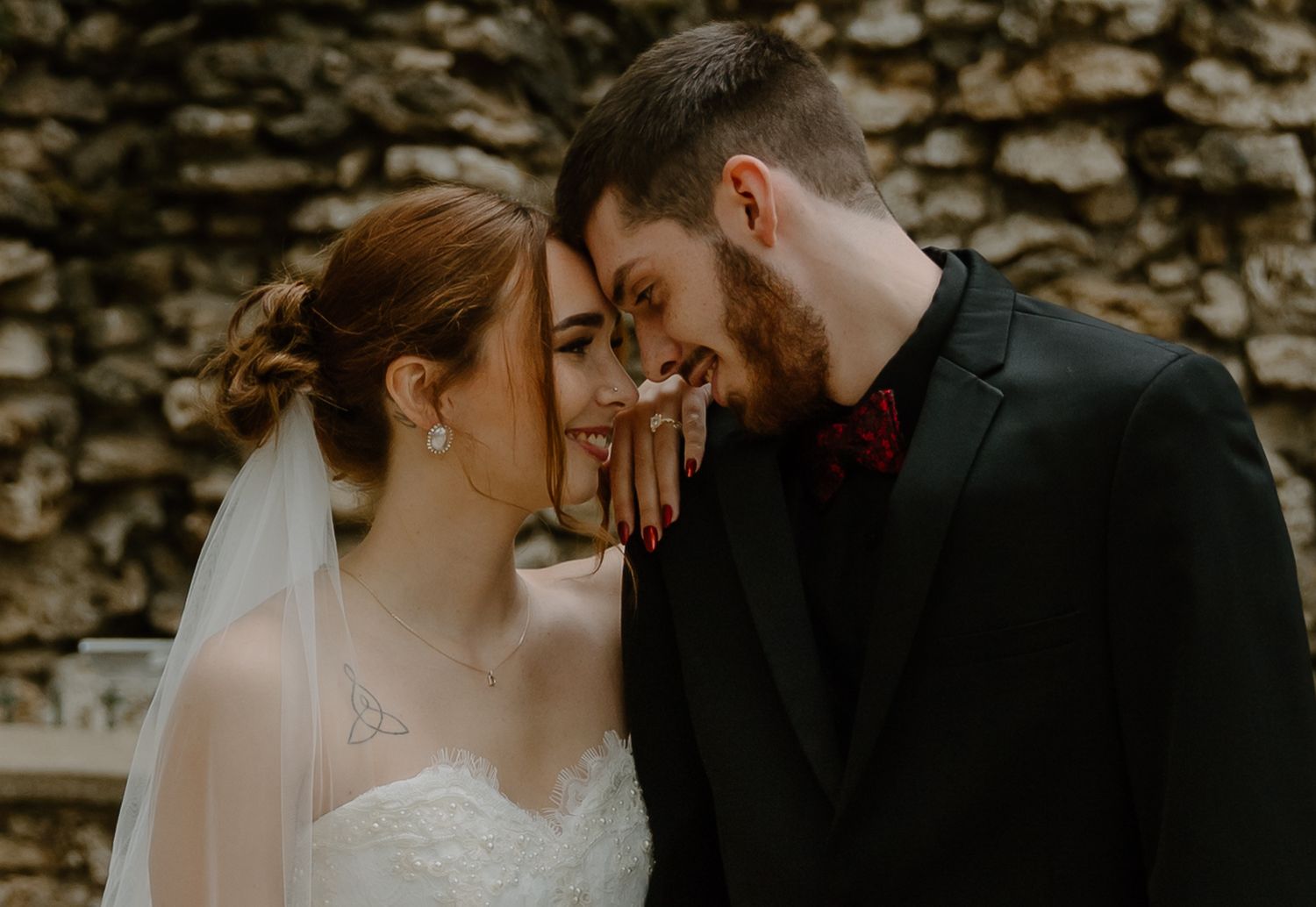 A romantic wedding couple shares an intimate moment against a rustic stone wall backdrop.