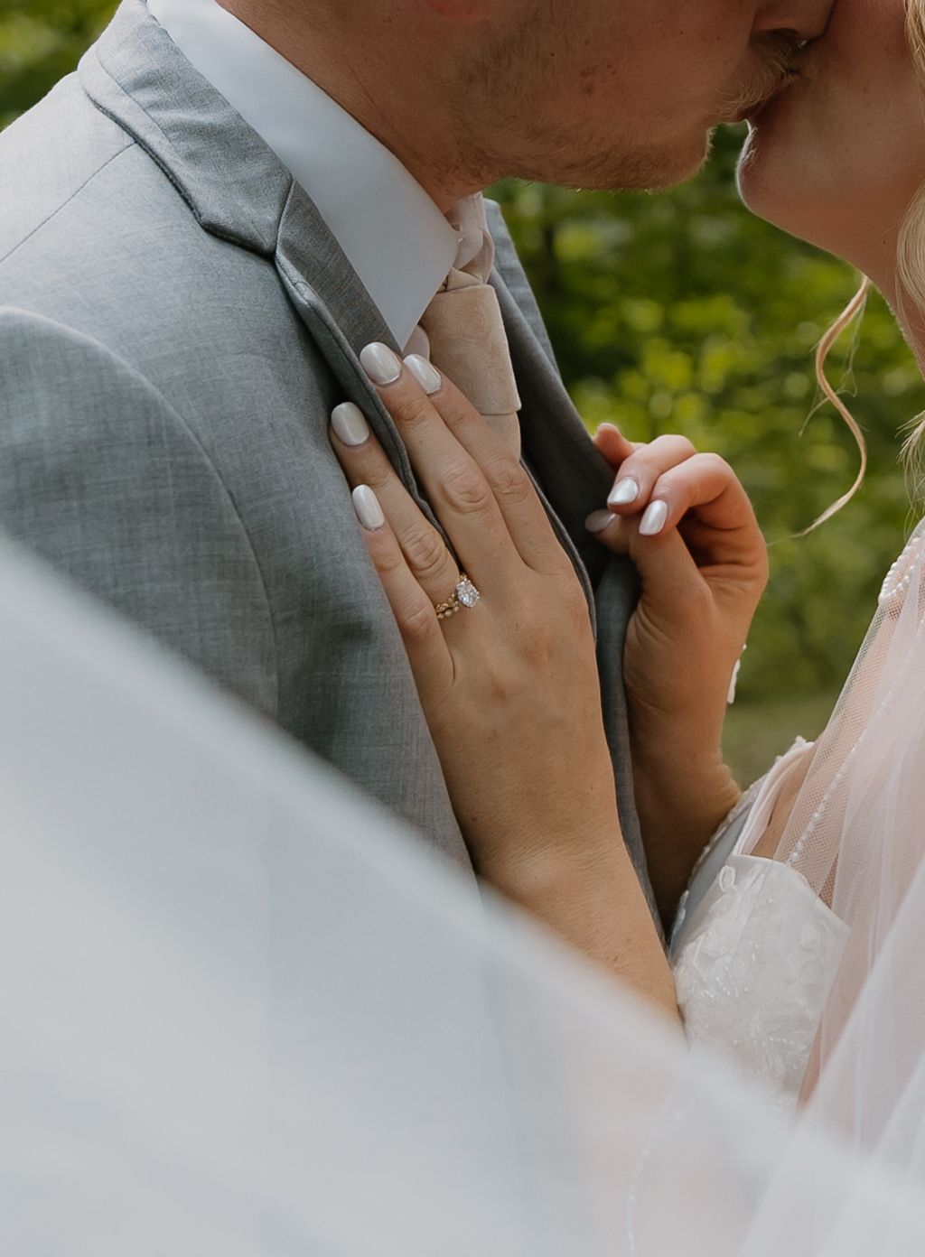 A bride and groom share a romantic kiss, her hand resting on his gray suit, adorned with a diamond engagement ring.
