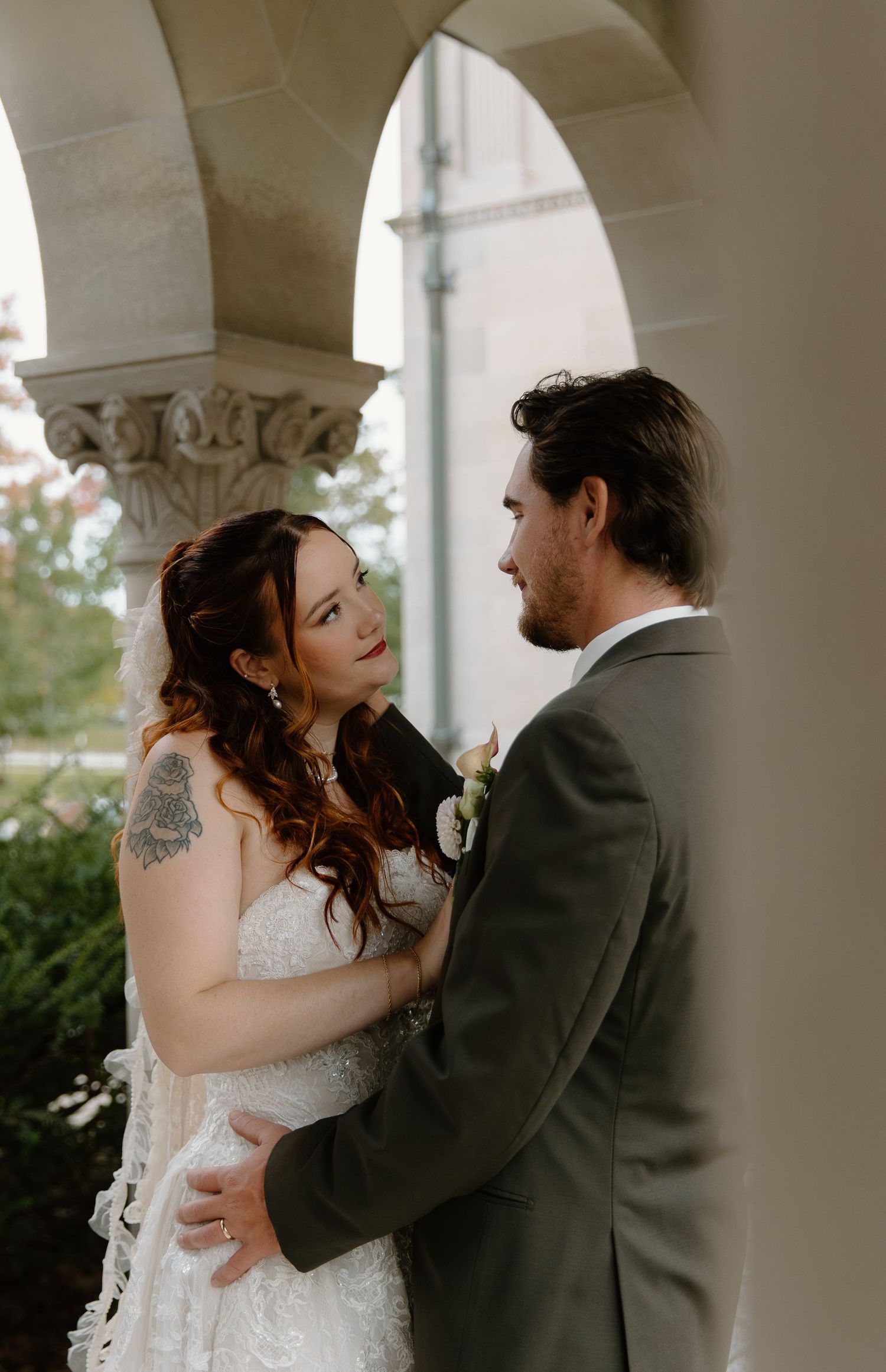A bride and groom share an intimate moment under stone arches, the bride in lace with a veil and the groom in a gray suit.