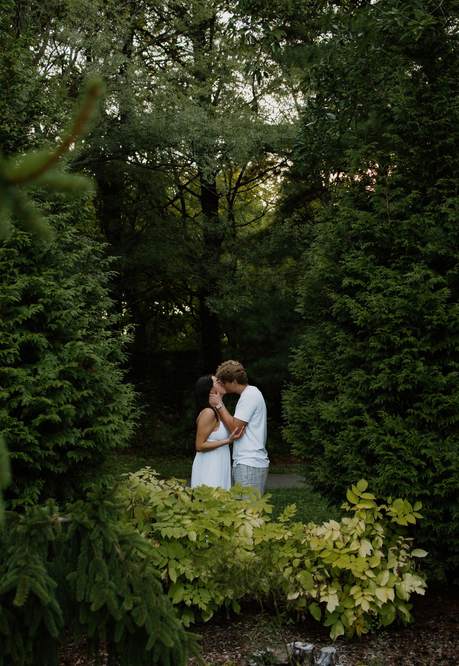 A couple shares a romantic moment surrounded by lush green trees and foliage in a serene garden setting.