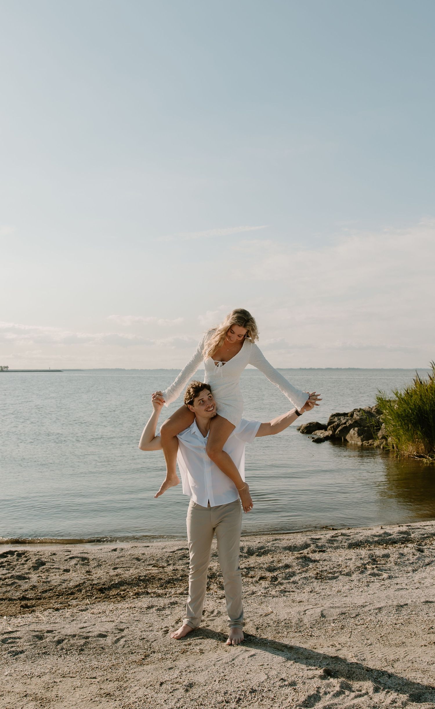 A couple enjoys a sunny beach day, with a woman sitting on a man's shoulders near the water's edge.