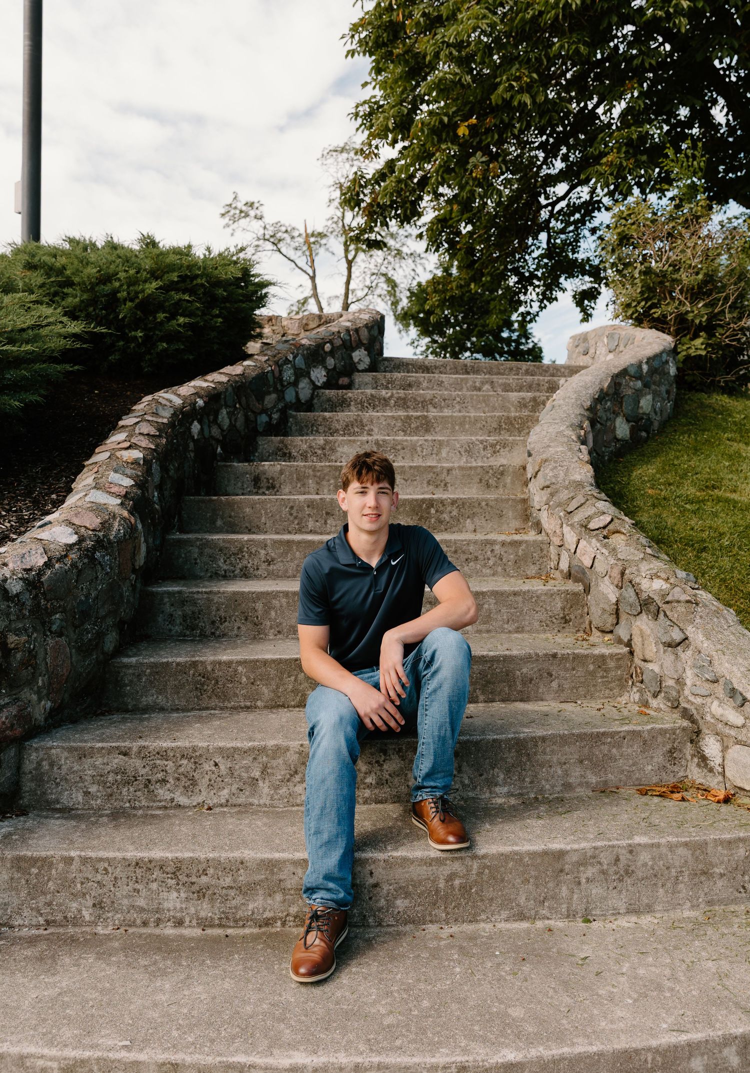 Young man in navy polo and jeans sitting on curved stone staircase outdoors.