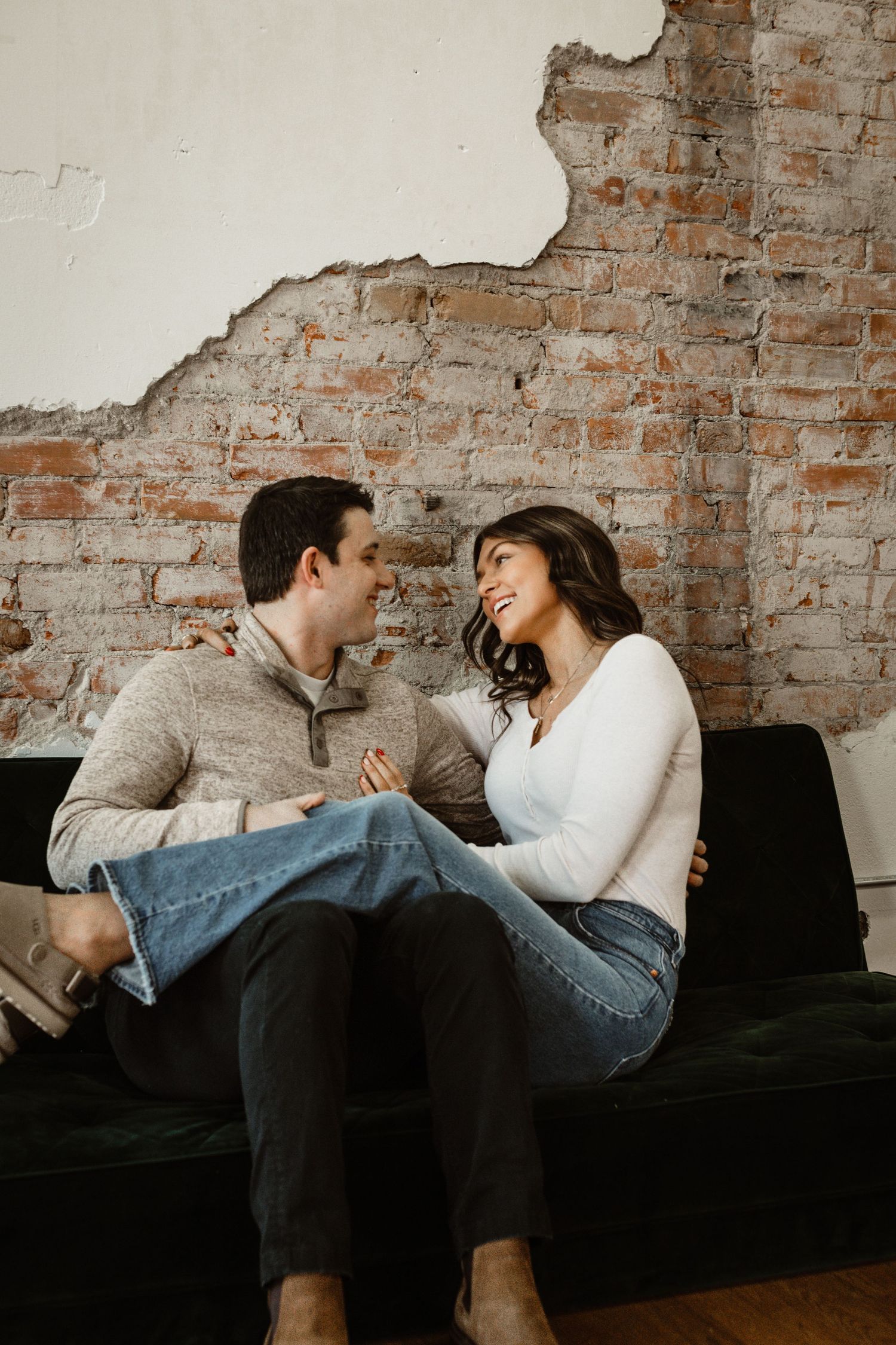 A couple laughs together while sitting on a black couch against a brick wall.