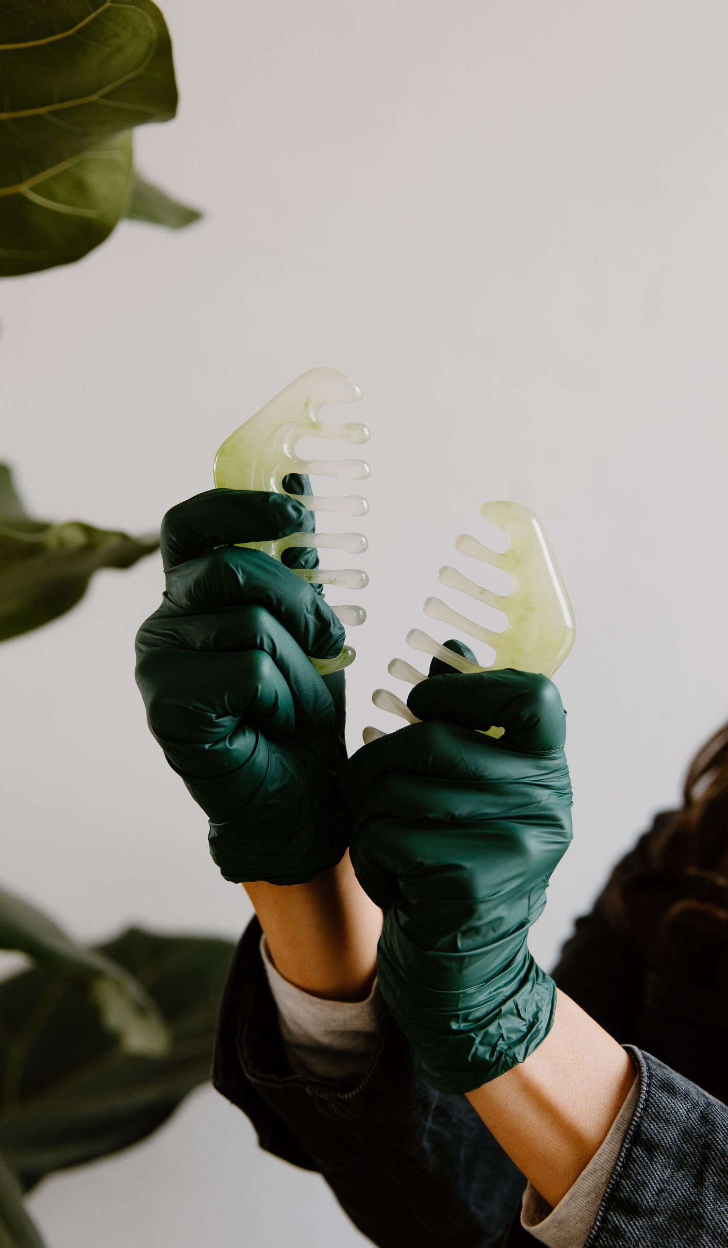 Hands in dark green latex gloves holding two jade gua sha combs against a light background with green leaves.