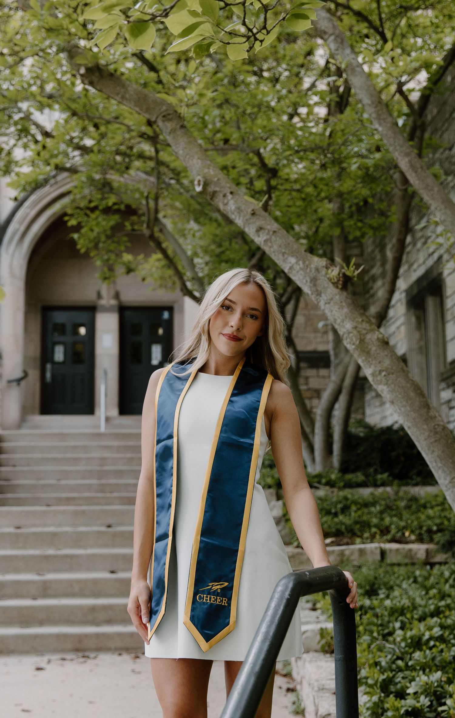 A smiling graduate poses in a white dress with a navy and gold cheer stole on stone steps surrounded by trees.