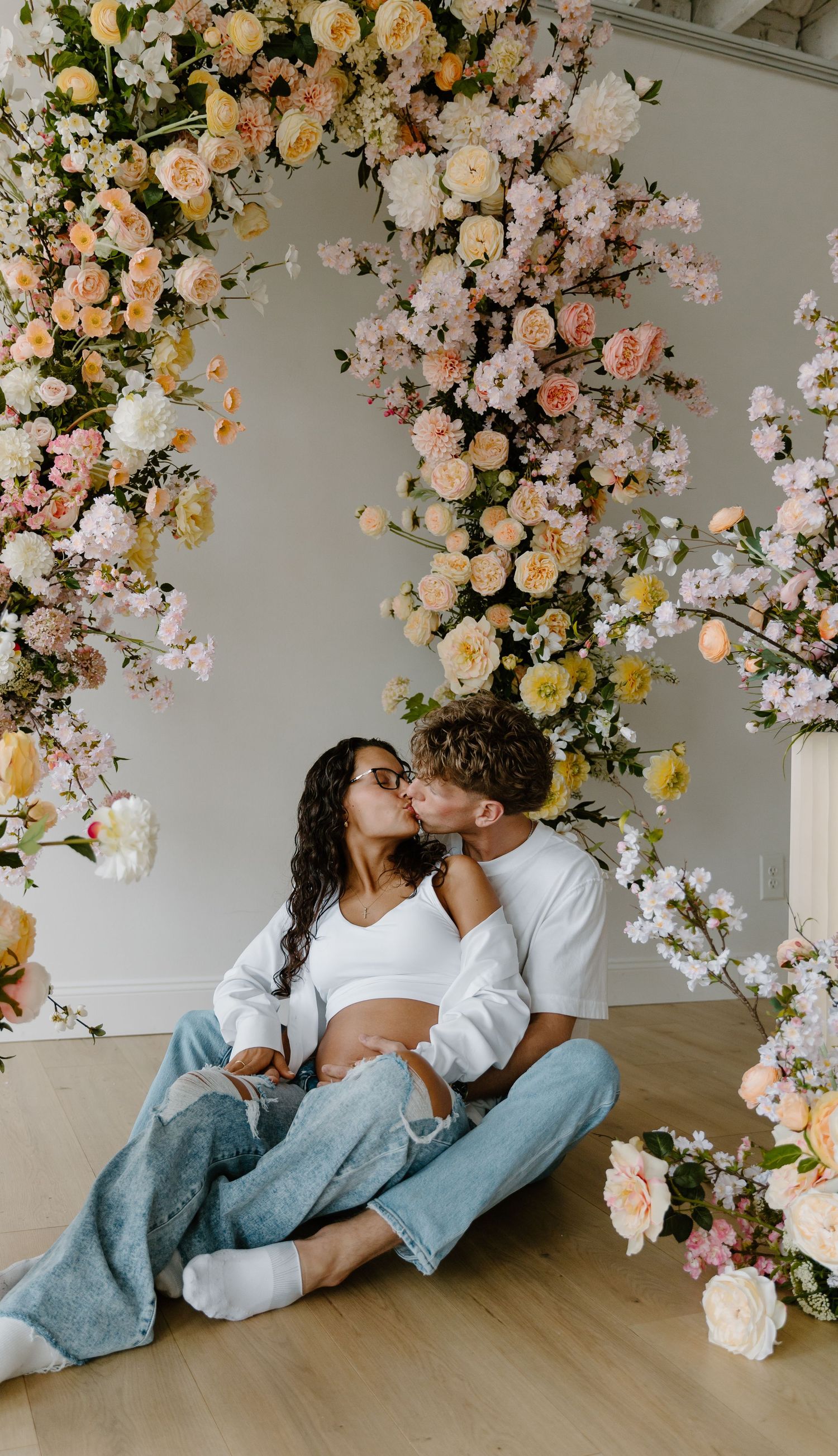 Expecting couple sharing a kiss while sitting on the floor surrounded by a beautiful pastel floral arrangement.