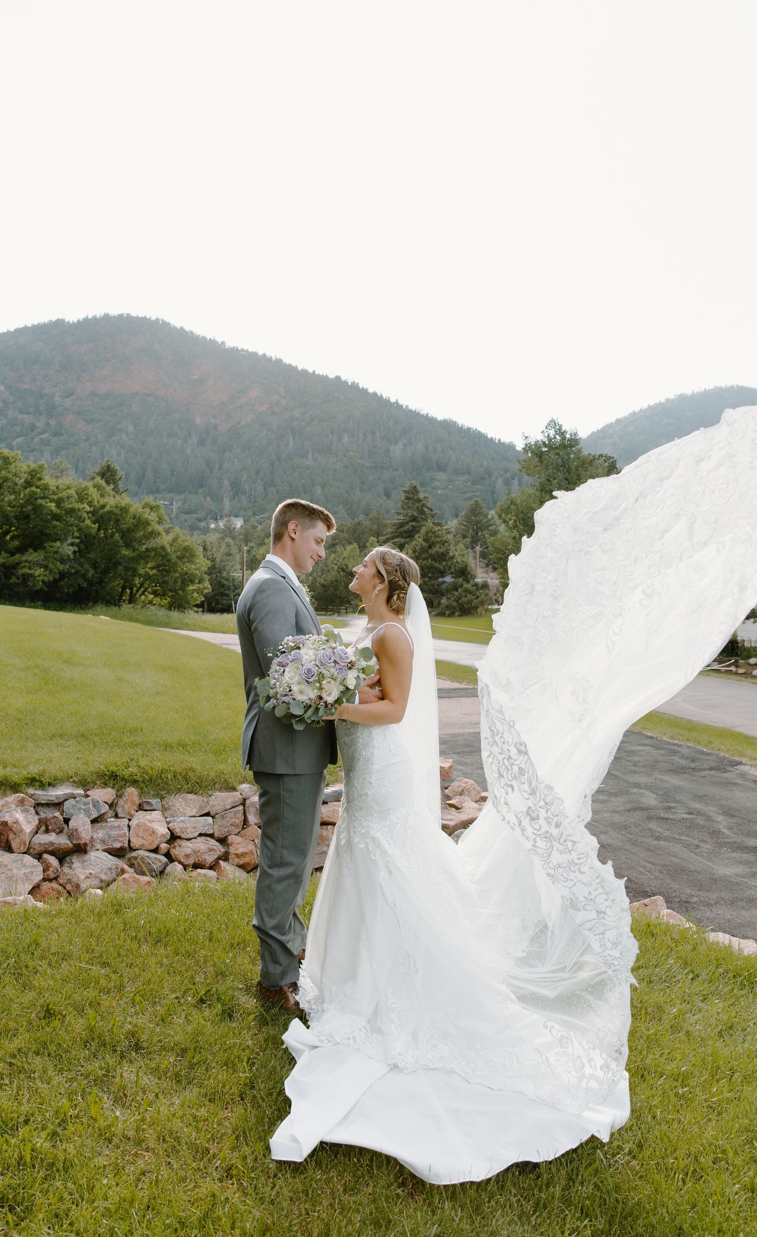 A couple shares an intimate moment on their wedding day as the bride's white dress flows in the mountain breeze.