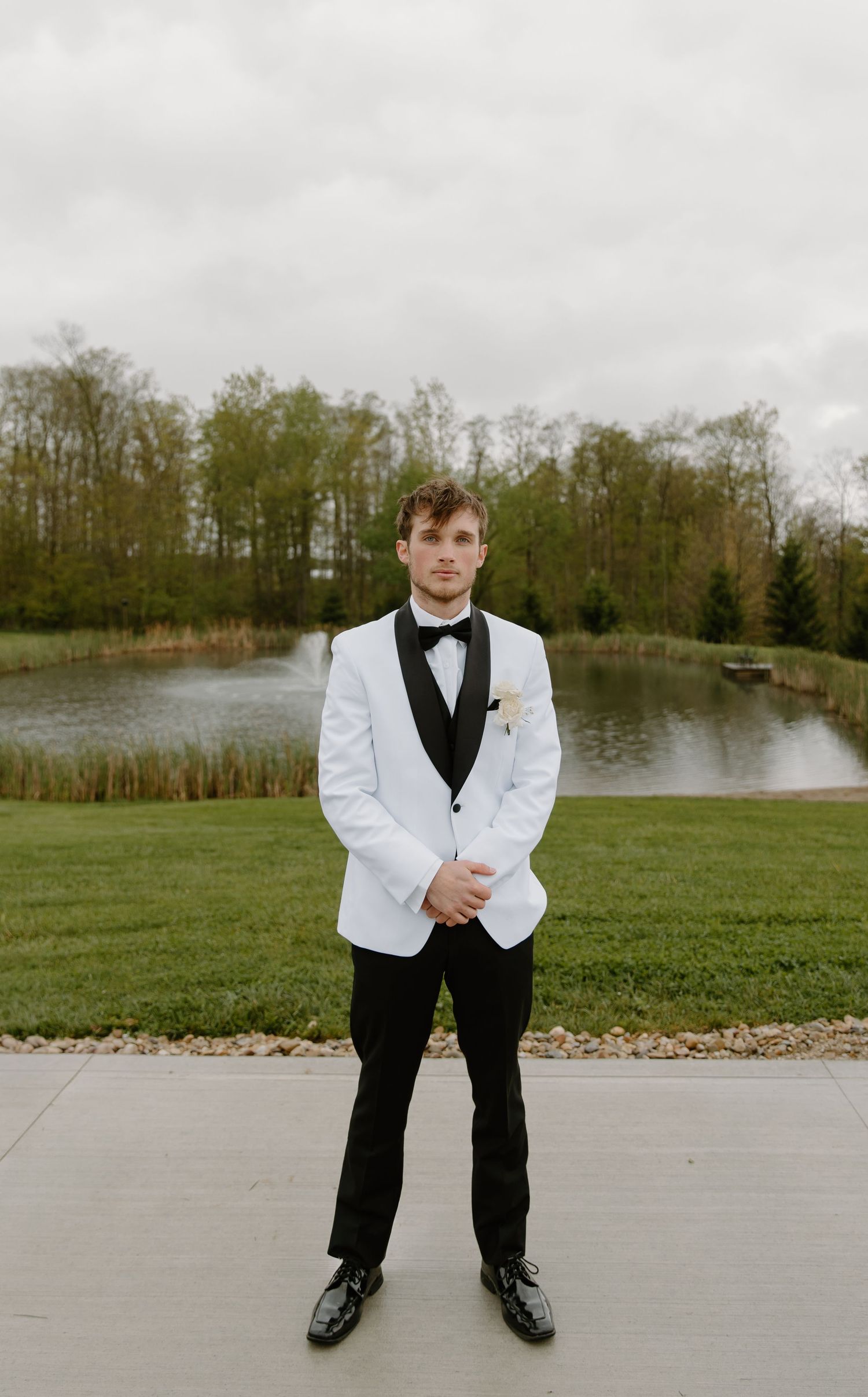 Groom in white tuxedo jacket and black bow tie standing confidently outdoors near a serene pond.