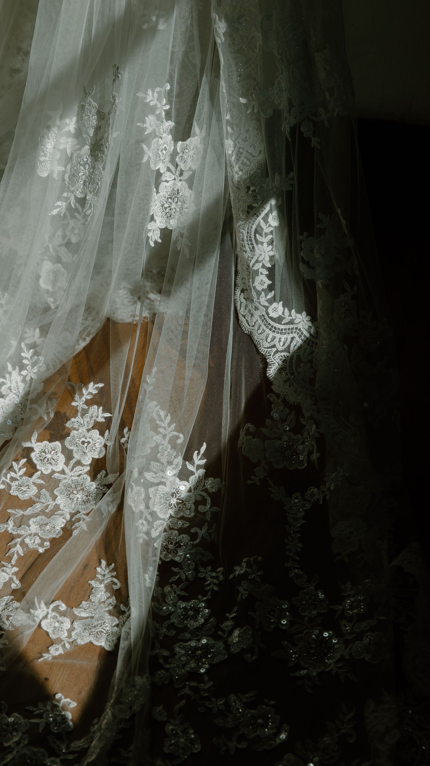Delicate white lace curtains with floral embroidery patterns filter soft light through a dark window.