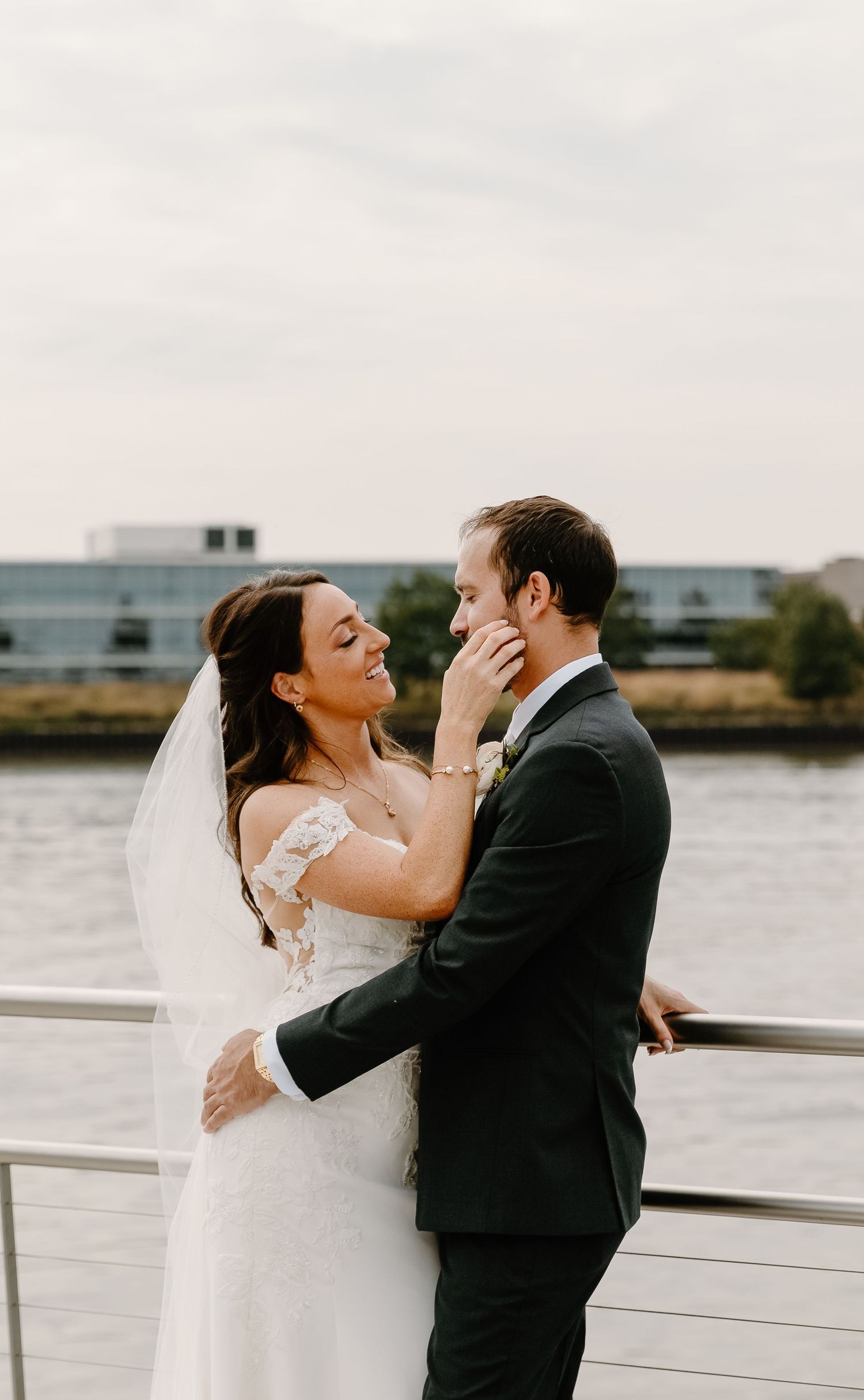 A romantic wedding couple embraces on a waterfront deck with city buildings in the background.