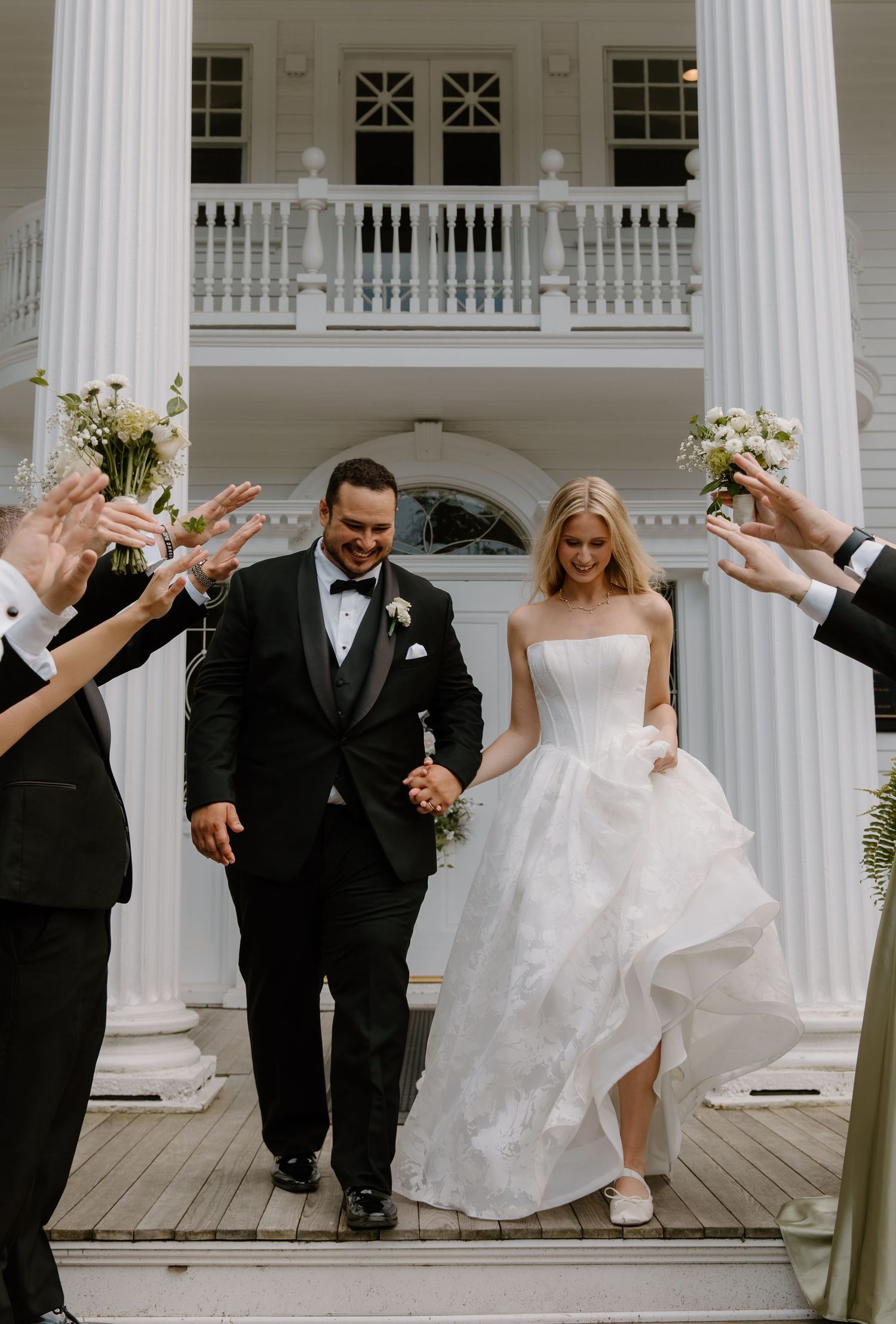 Bride and groom exit a white colonial mansion as guests celebrate their wedding ceremony.