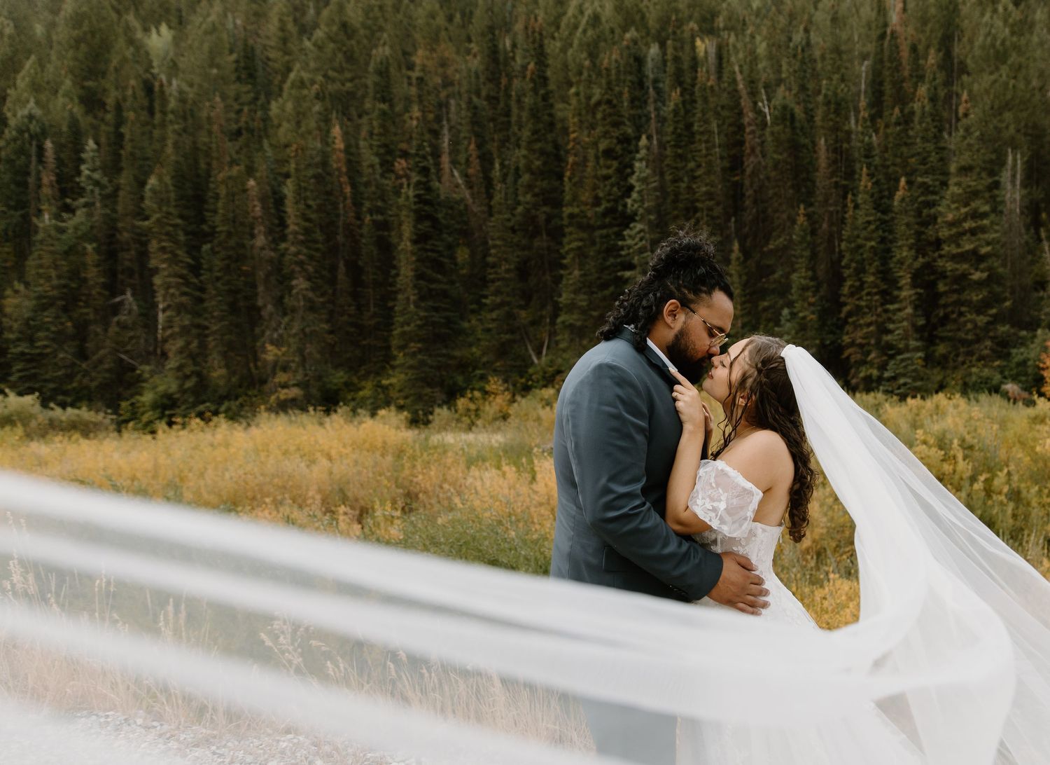 A couple shares a romantic kiss in a scenic mountain meadow with evergreen forest in the background.