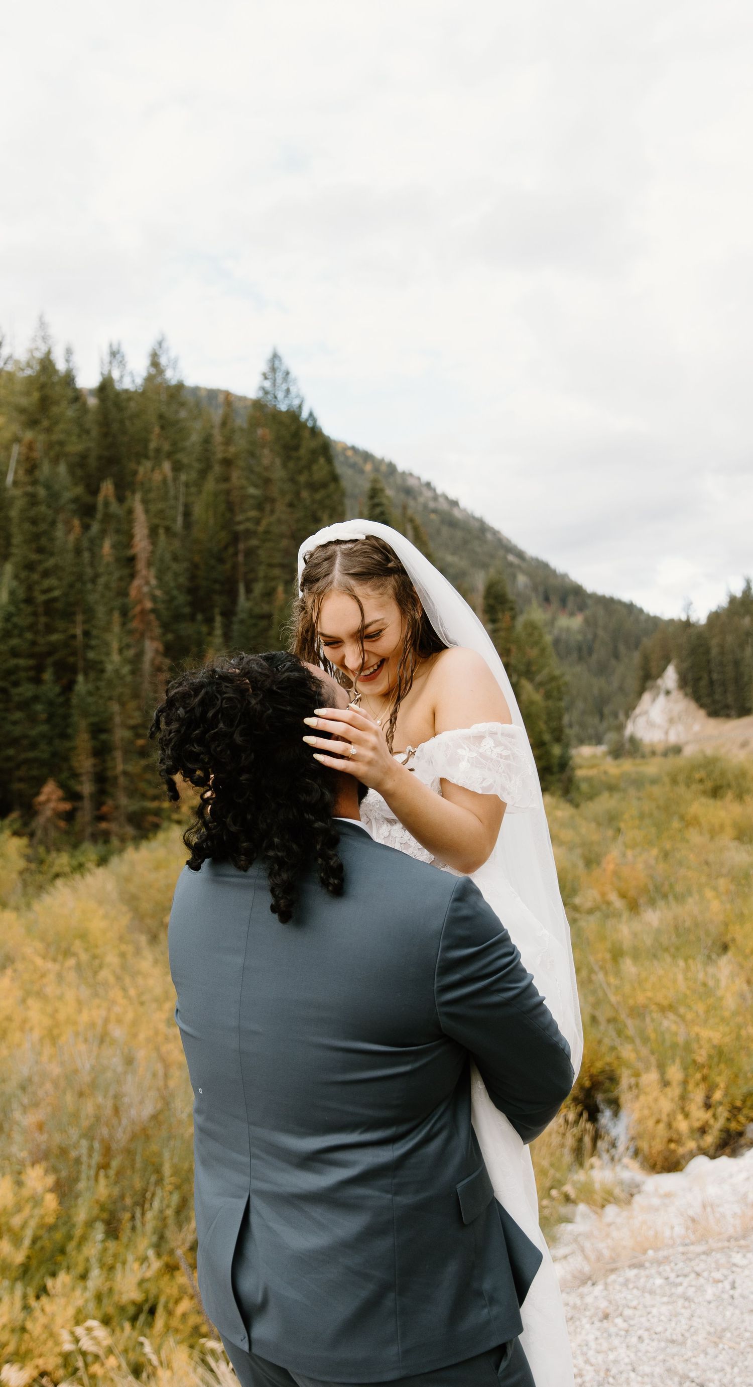 A romantic wedding couple embraces in a scenic mountain meadow surrounded by evergreen trees and autumn foliage.