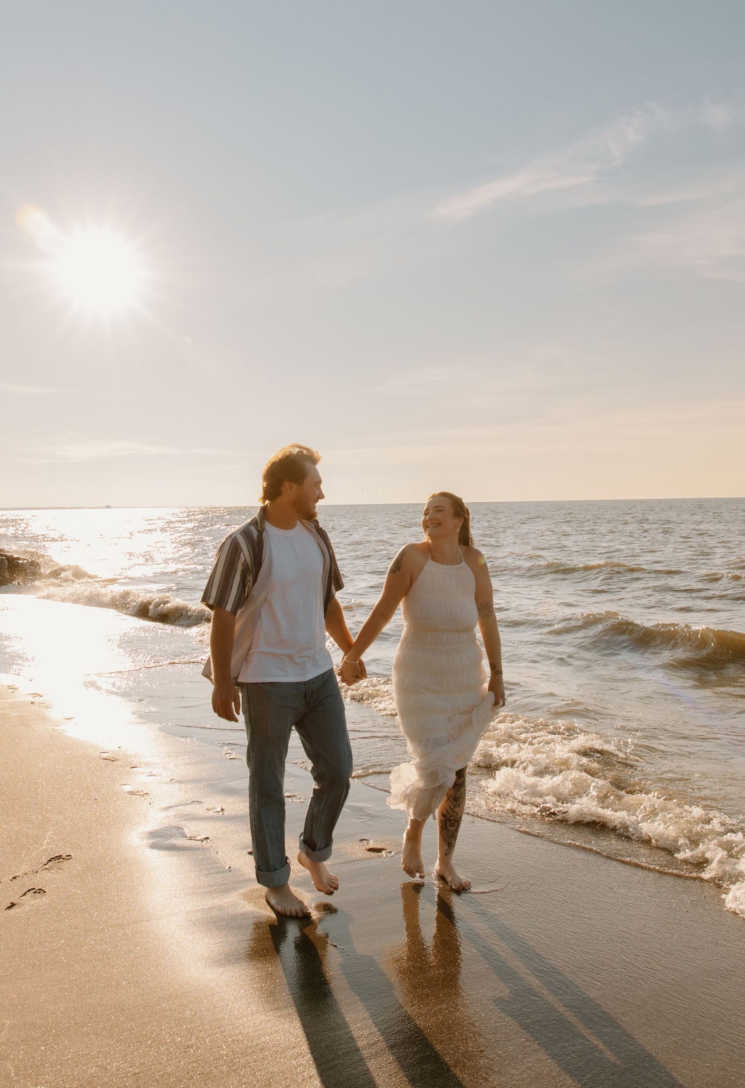 Couple holding hands walking barefoot on a sunny beach at golden hour, waves gently washing ashore.