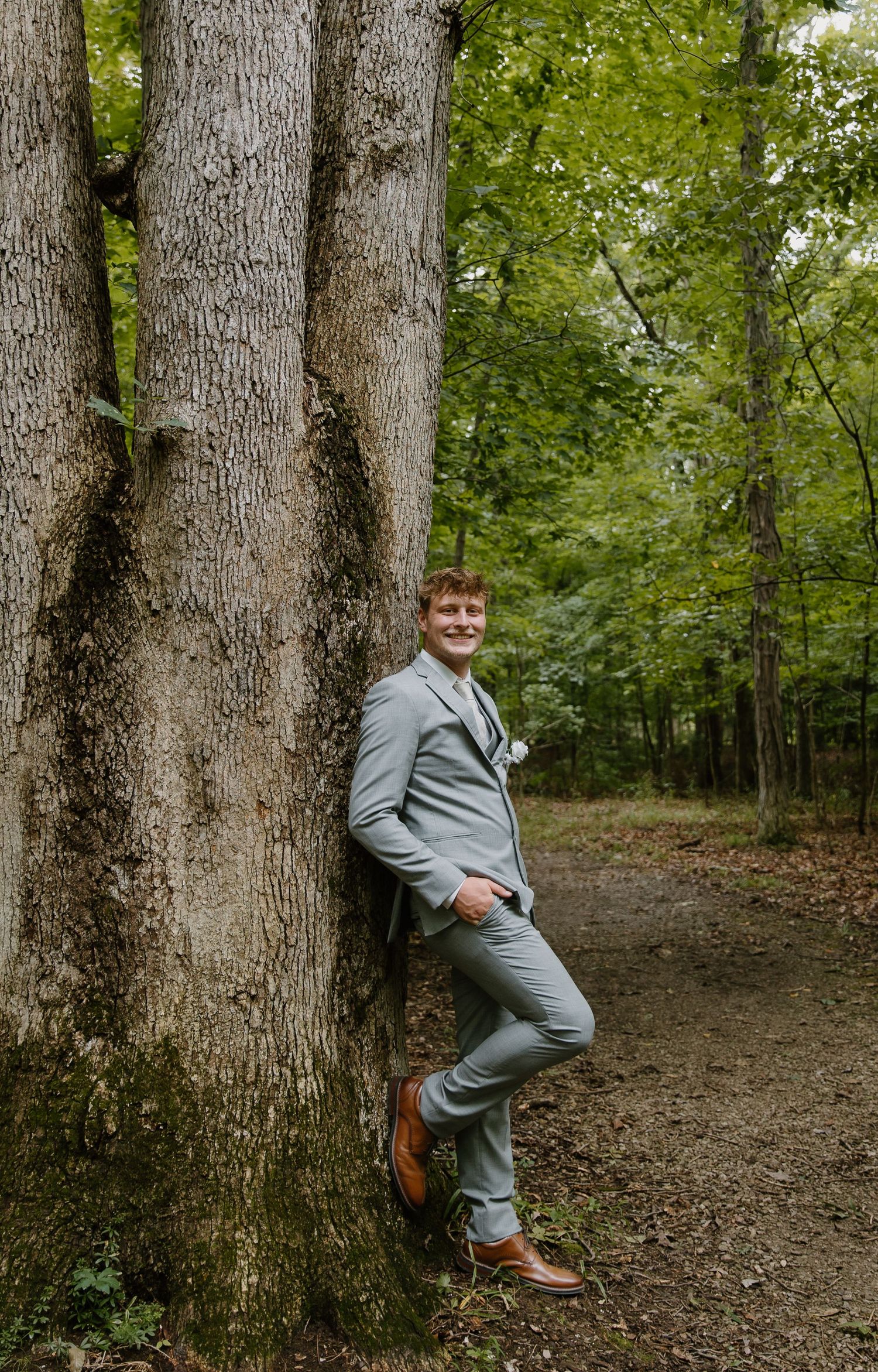 Young man in gray suit leaning against tree trunk in forest, smiling confidently for formal portrait photo.