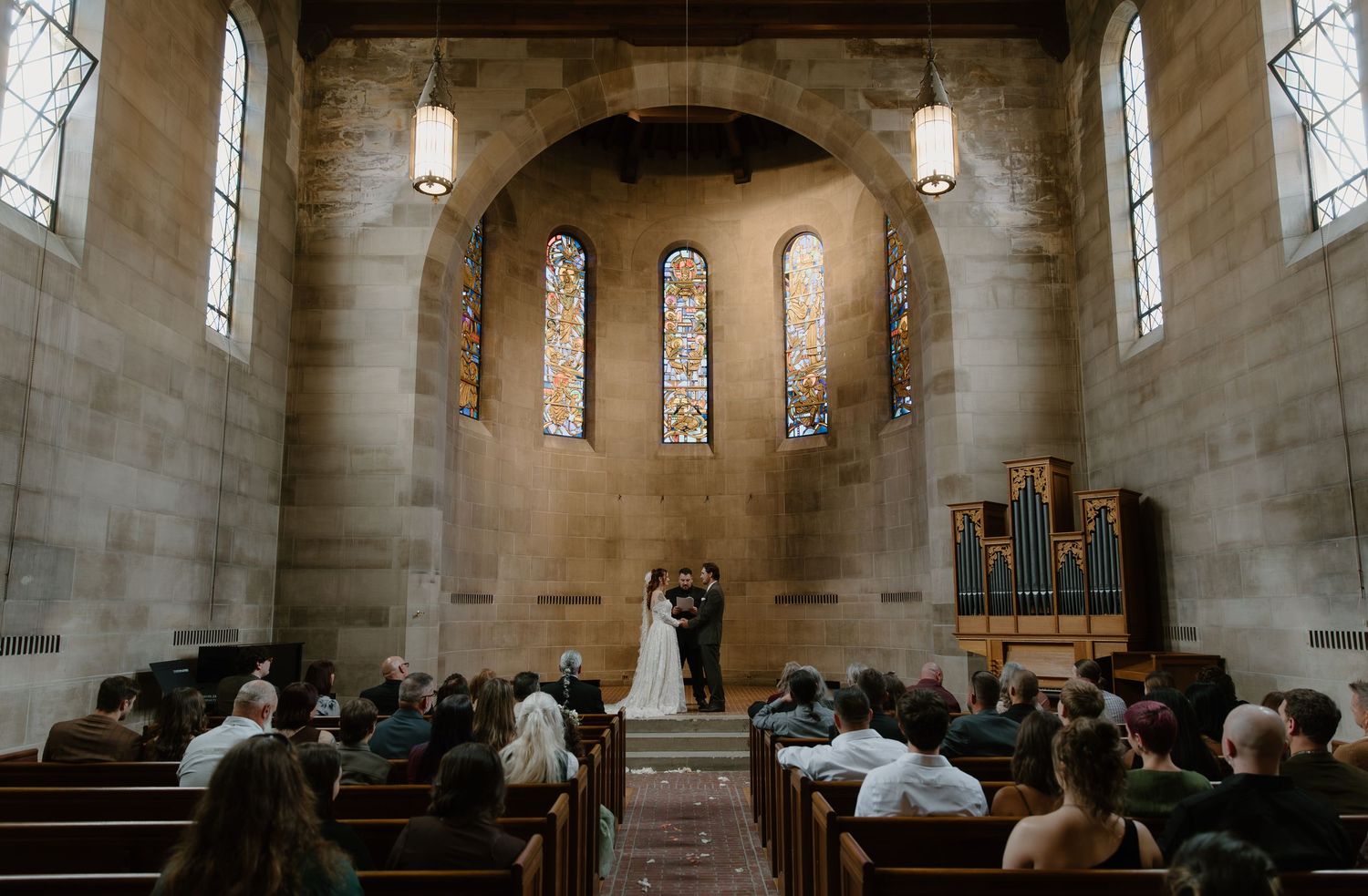Wedding ceremony in a stone church with stained glass windows, wooden pews, and an organ backdrop.