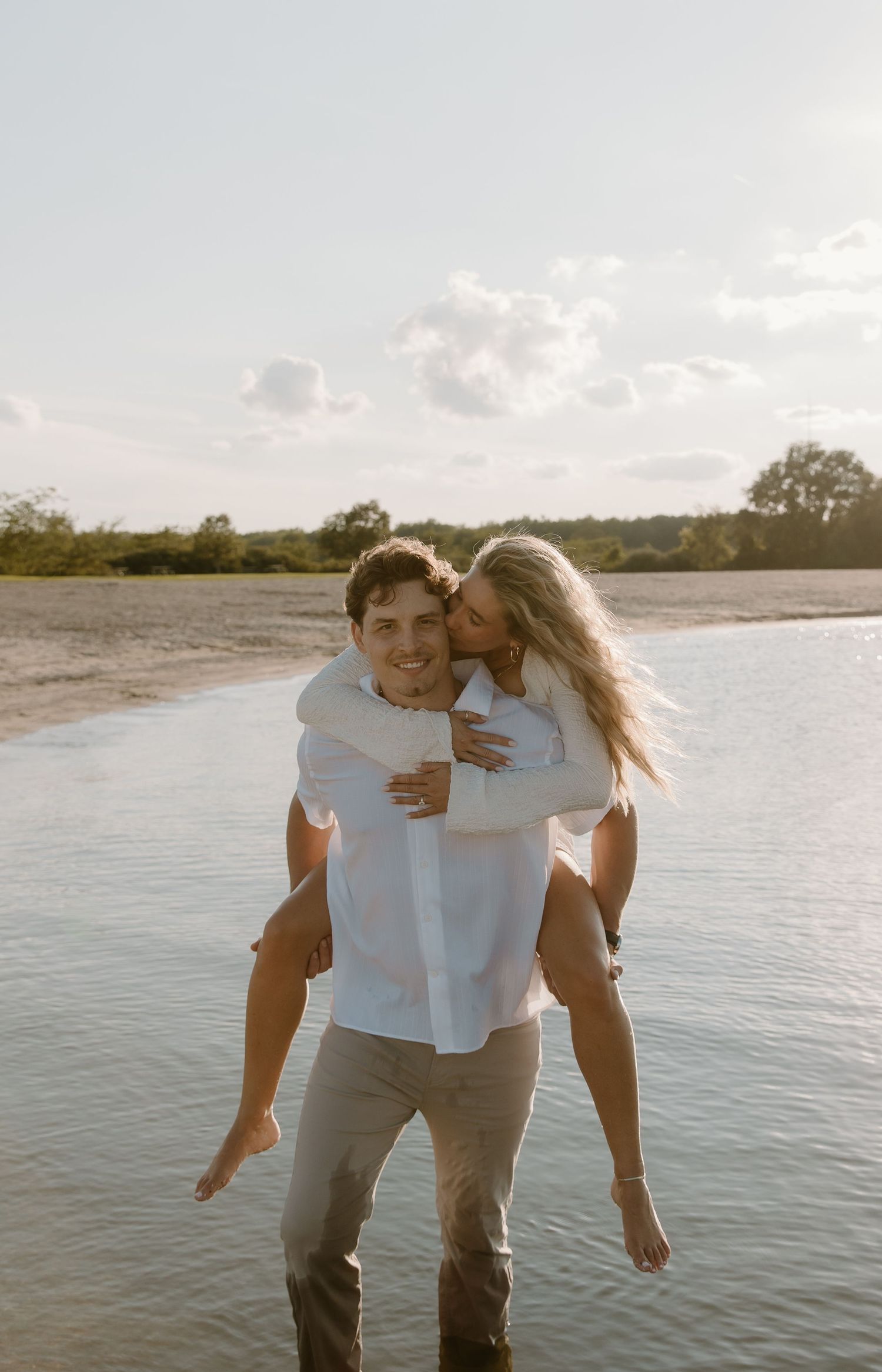 A smiling man gives a piggyback ride to a blonde woman kissing his cheek while wading in shallow water outdoors.