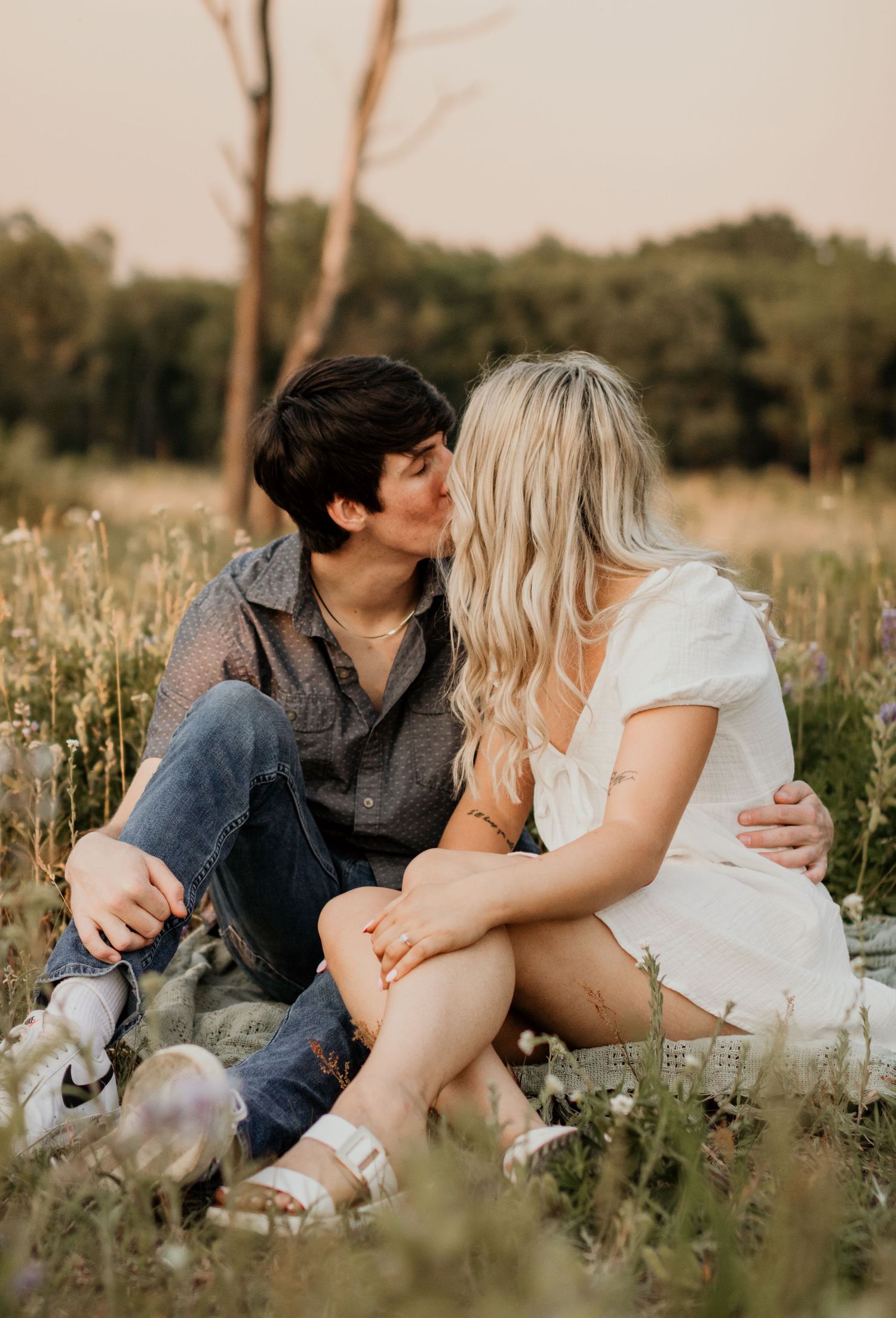 A couple sits close together in a meadow during a warm summer evening.