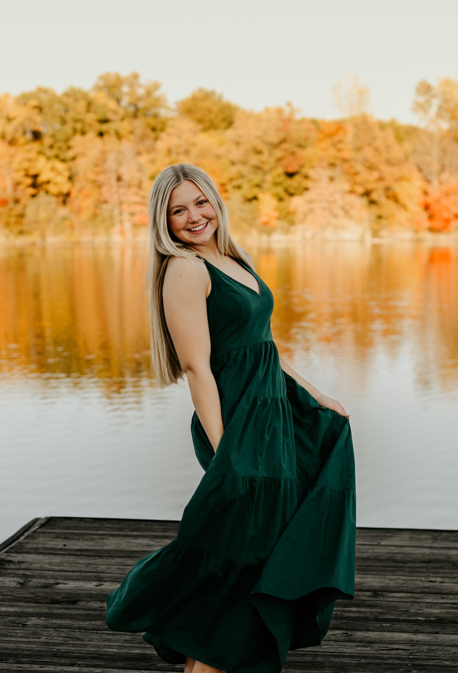Woman in emerald velvet dress twirling on wooden dock with autumn colors reflecting in lake.