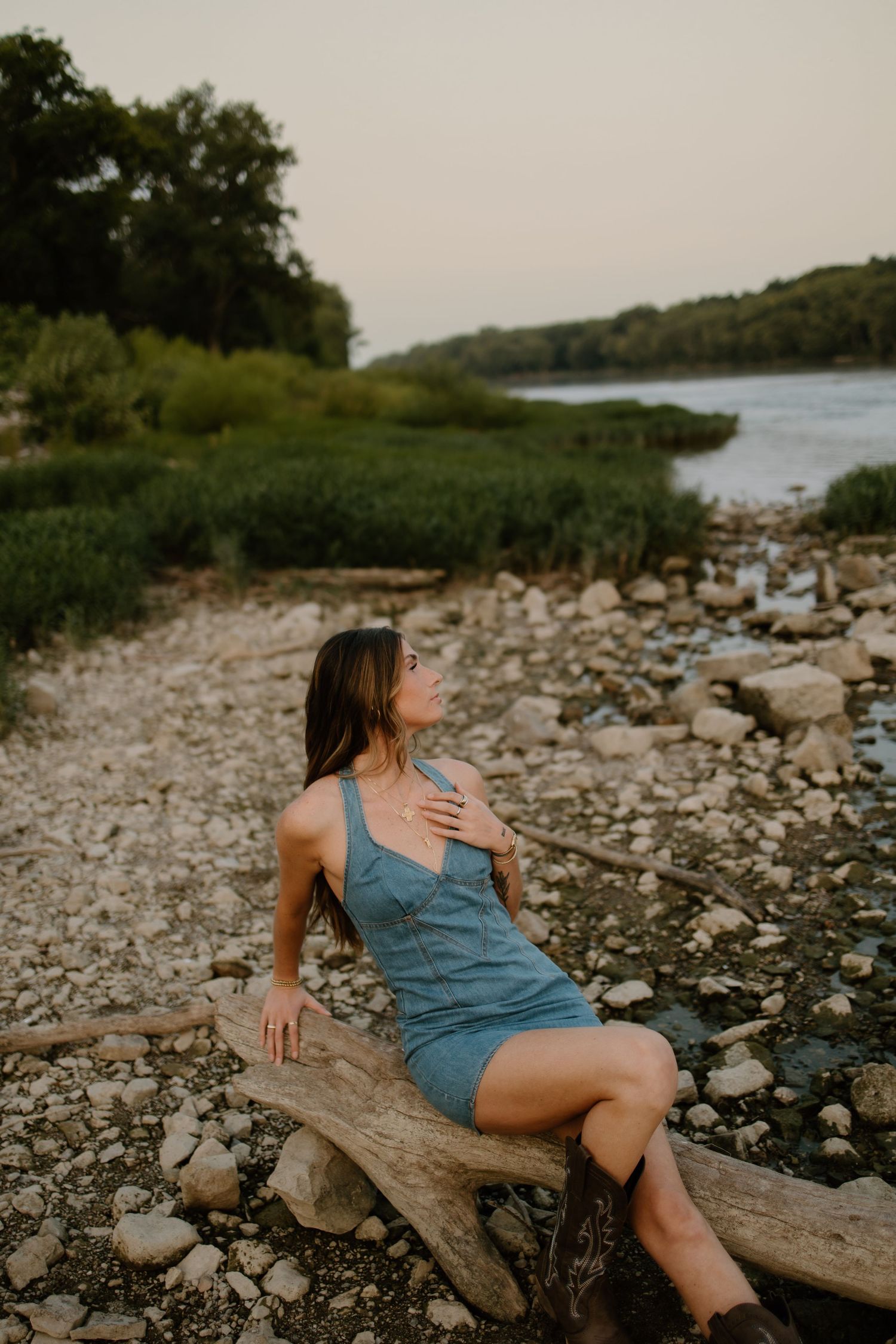 Woman in blue dress sitting on driftwood by rocky riverbank at sunset with lush green trees behind her.