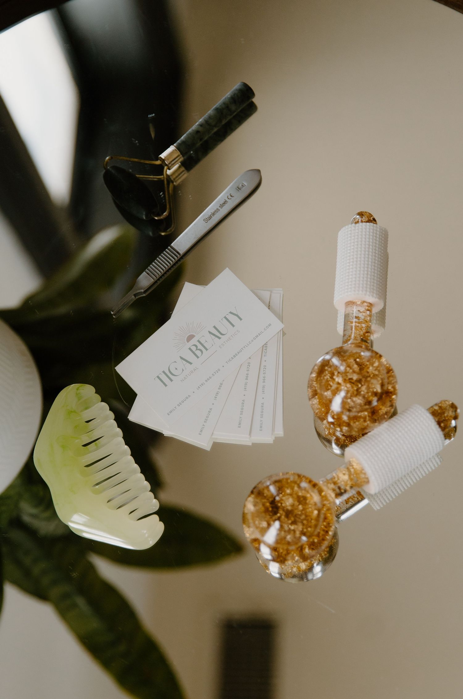 Flat lay of TIGA Beauty skincare tools including jade gua sha, face roller, serum bottles, and business cards on a neutral surface.