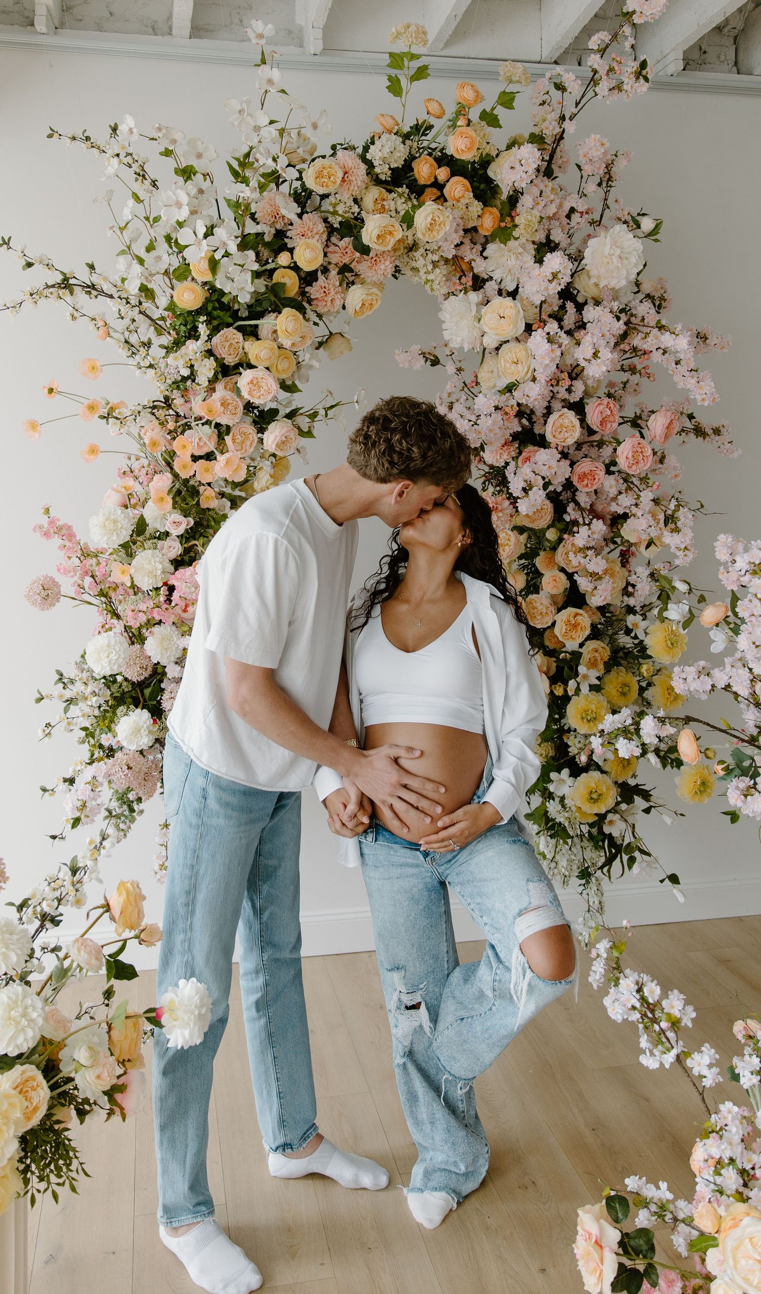 Pregnant couple kissing in front of a lush floral arch with peach and pink roses, wearing matching white tops and jeans.