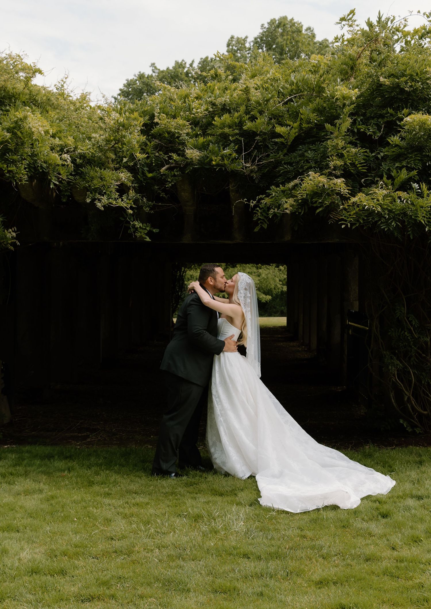 Bride and groom share a romantic kiss under a lush green pergola on their wedding day.