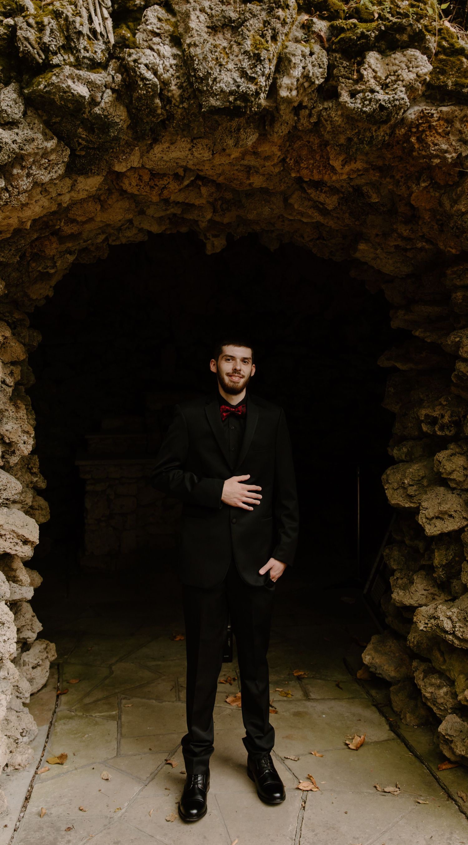 Black dress shoes and formal groom attire visible in a stone cave entrance during a wedding photoshoot.