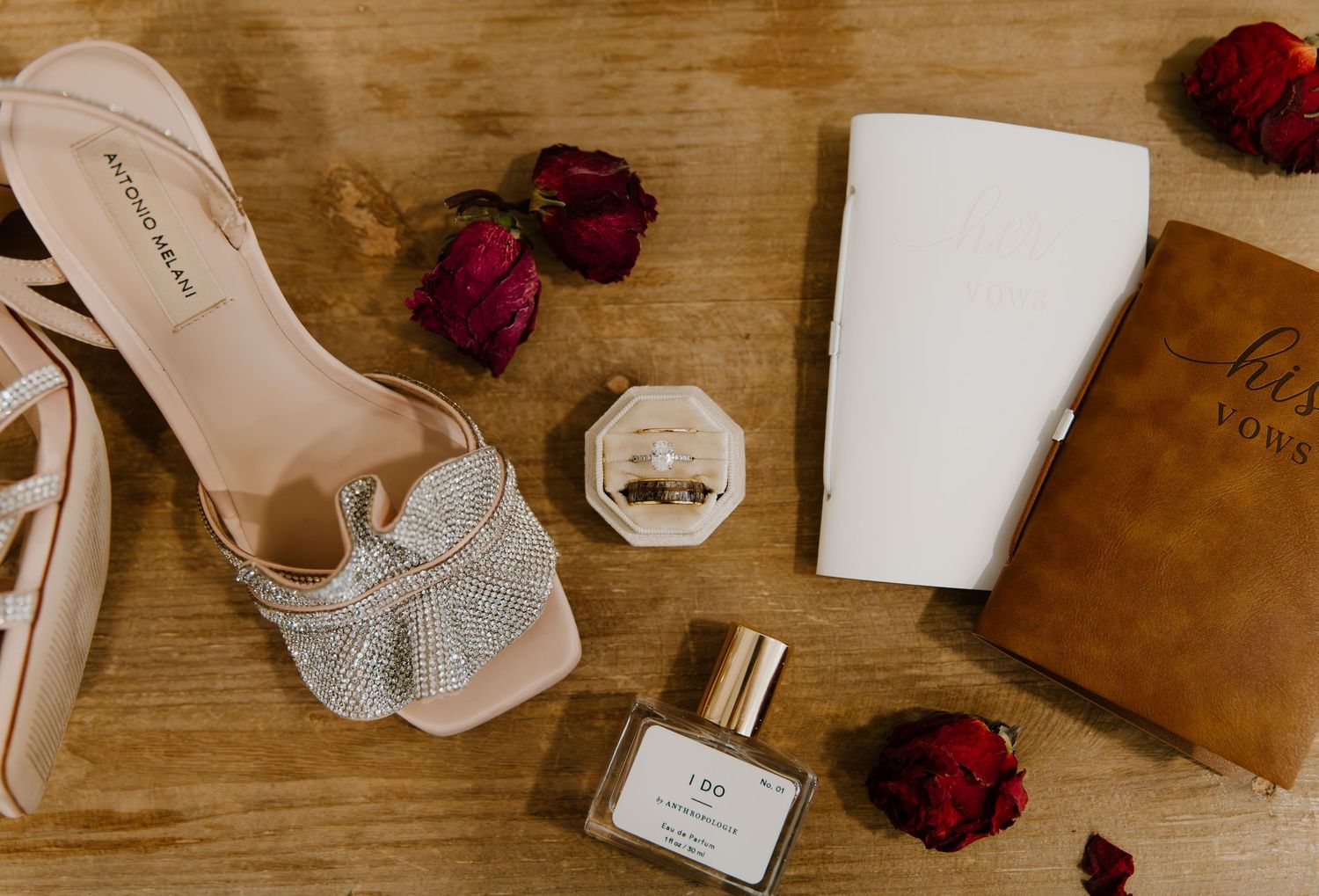Wedding accessories including bridal shoes, perfume, and dried roses arranged on rustic wooden surface.