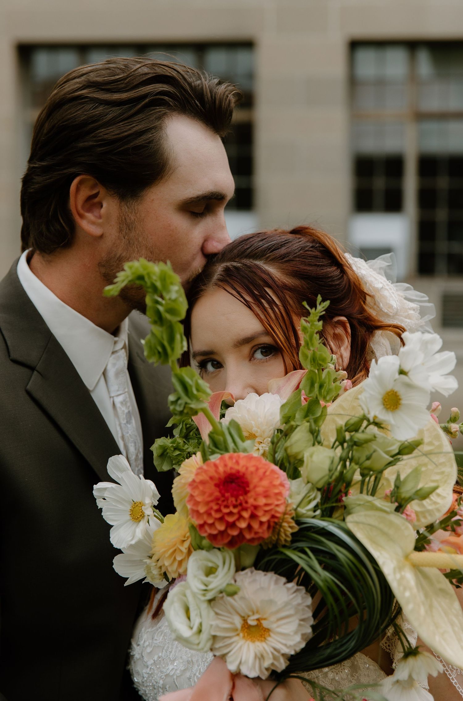 Groom kissing bride on head while she holds a lush bouquet of dahlias, cosmos, and greenery.
