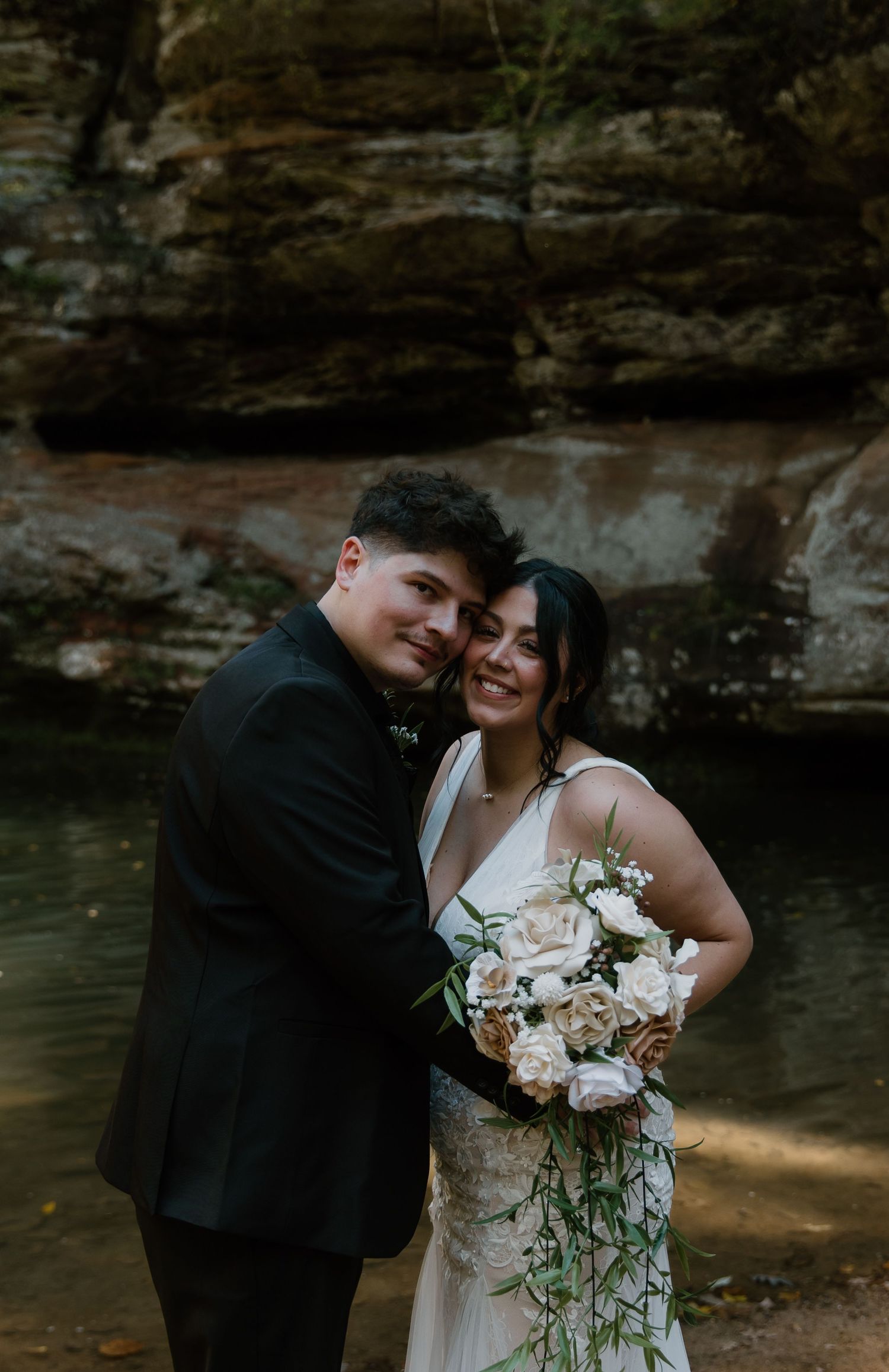 A smiling bride holding a cascading white rose bouquet poses with her groom in a black suit before a rocky waterfall backdrop.