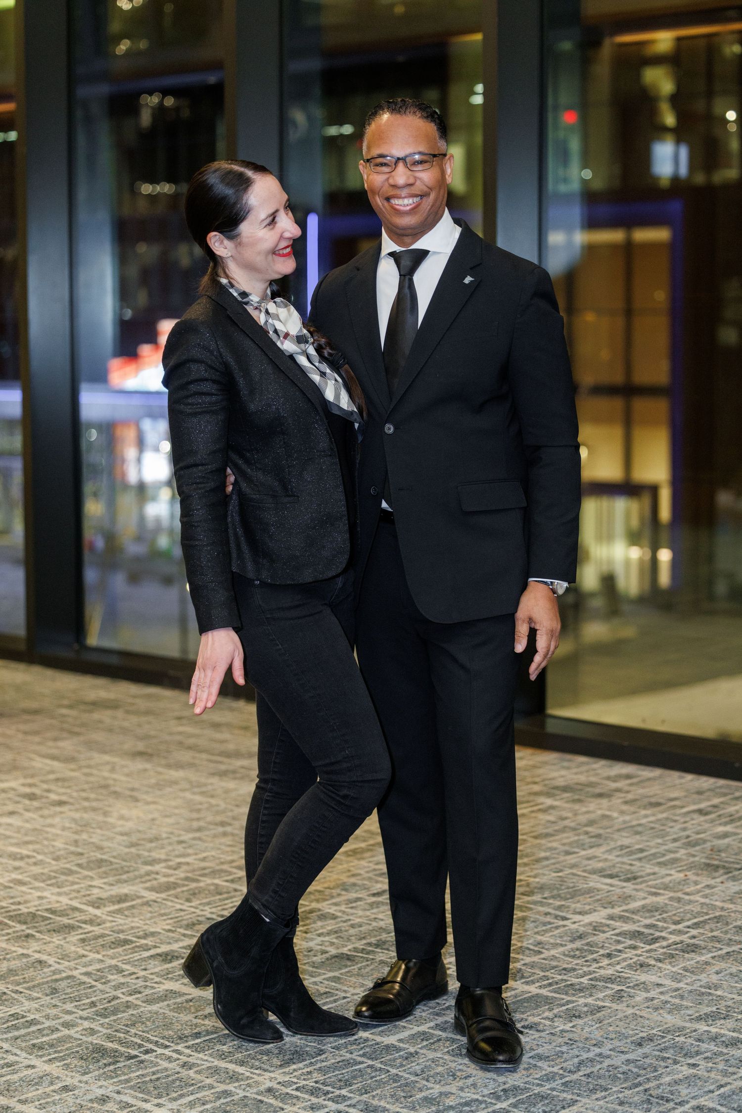 Two people in formal black attire share a joyful moment while walking together on a tiled floor.