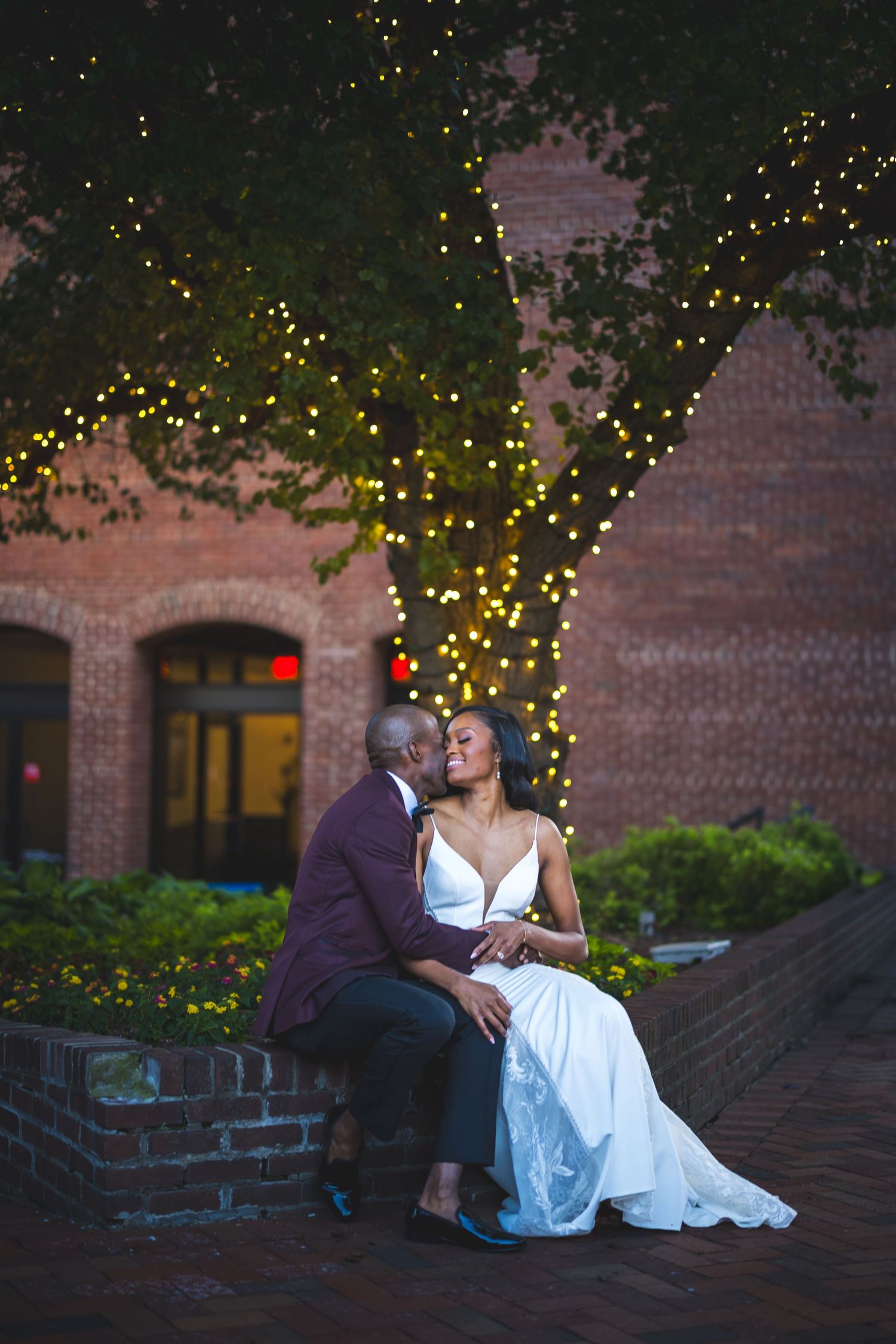 A couple shares an intimate moment seated on brick steps with twinkling string lights in the background at dusk.