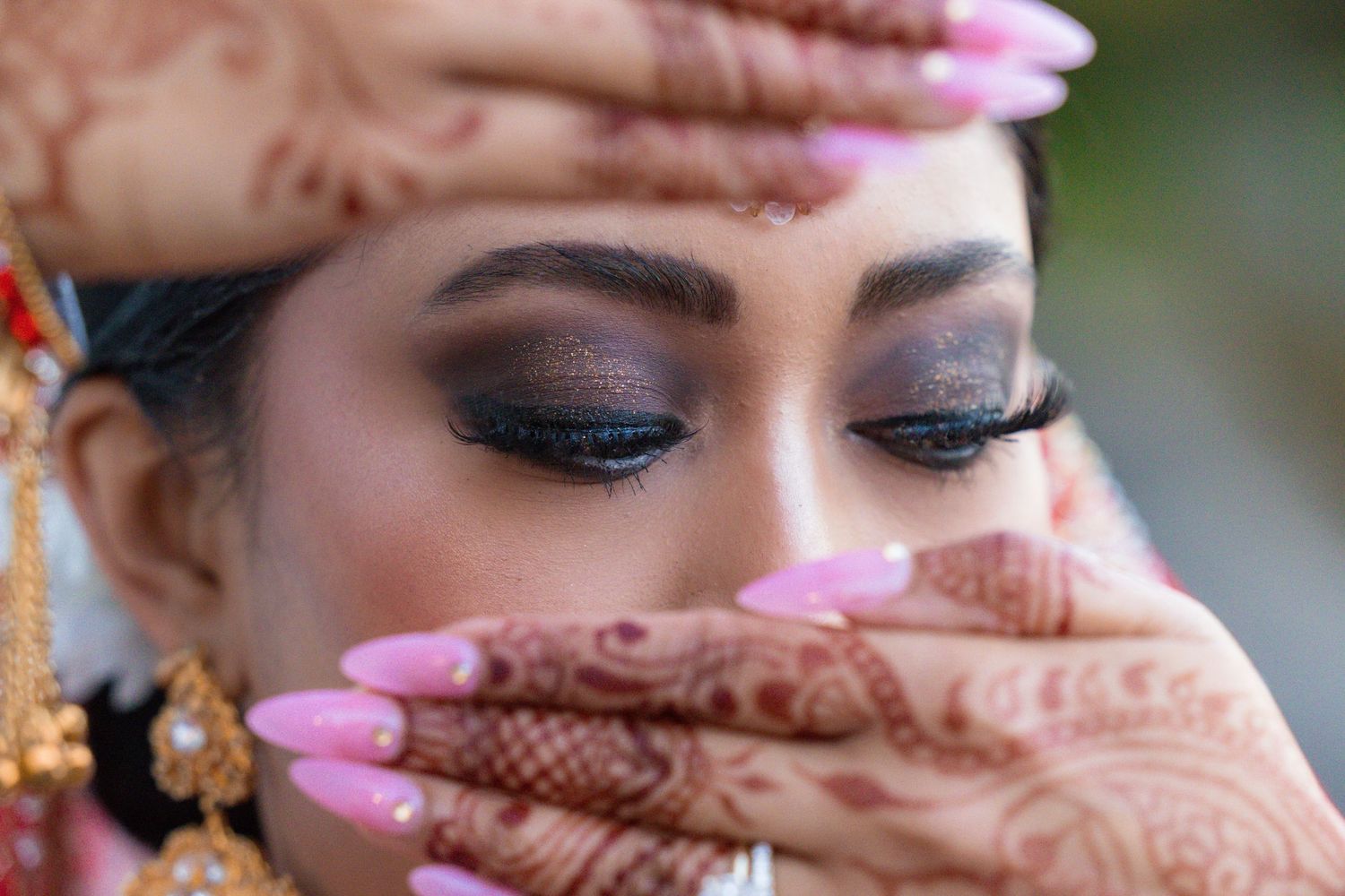 Close-up of traditional henna design on hand with pink nail polish and dramatic eye makeup visible in frame.