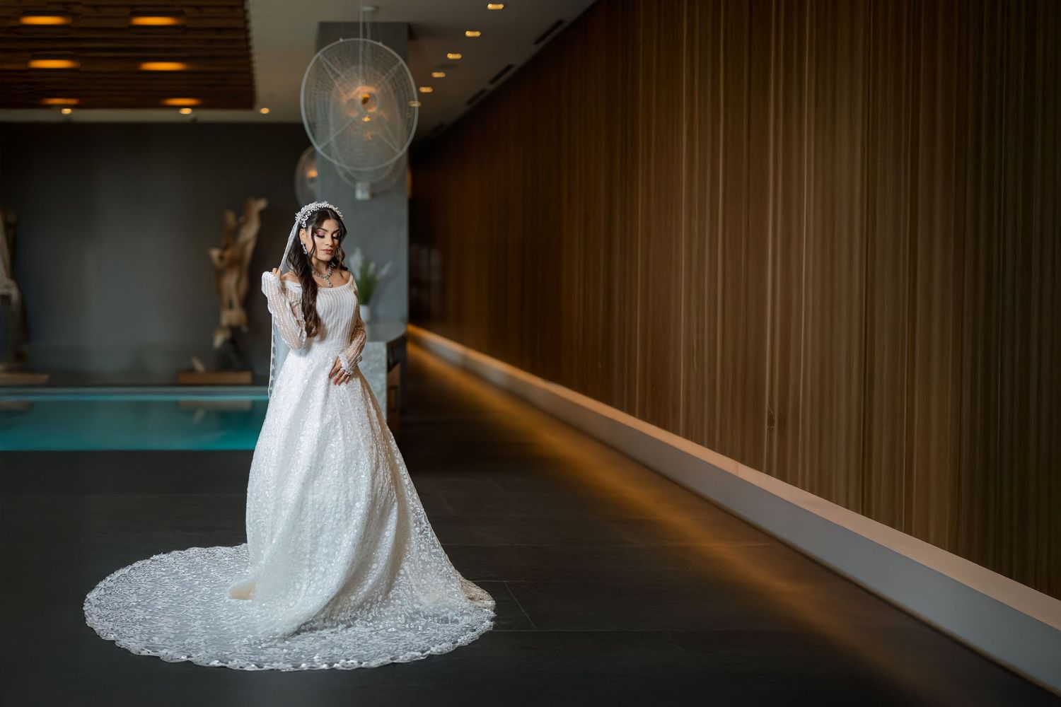 A bride in a lace wedding gown poses elegantly in a modern hallway with wood paneling and soft lighting.