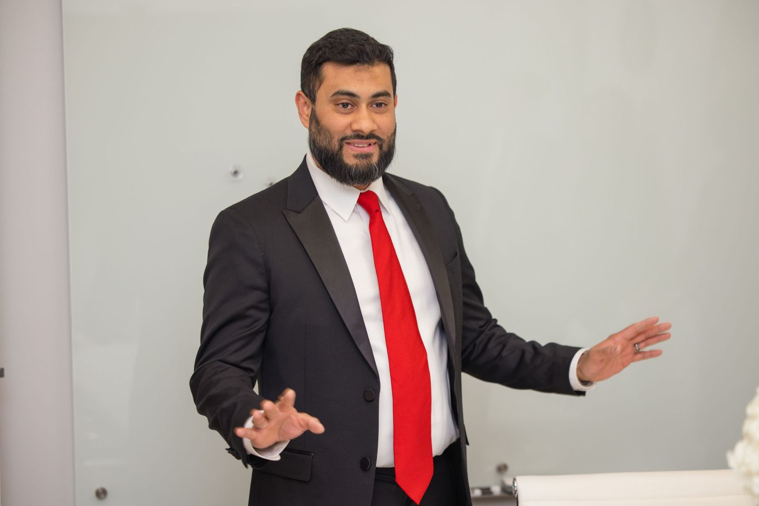 Professional in a black suit and red tie gestures expressively while presenting in a white office setting.